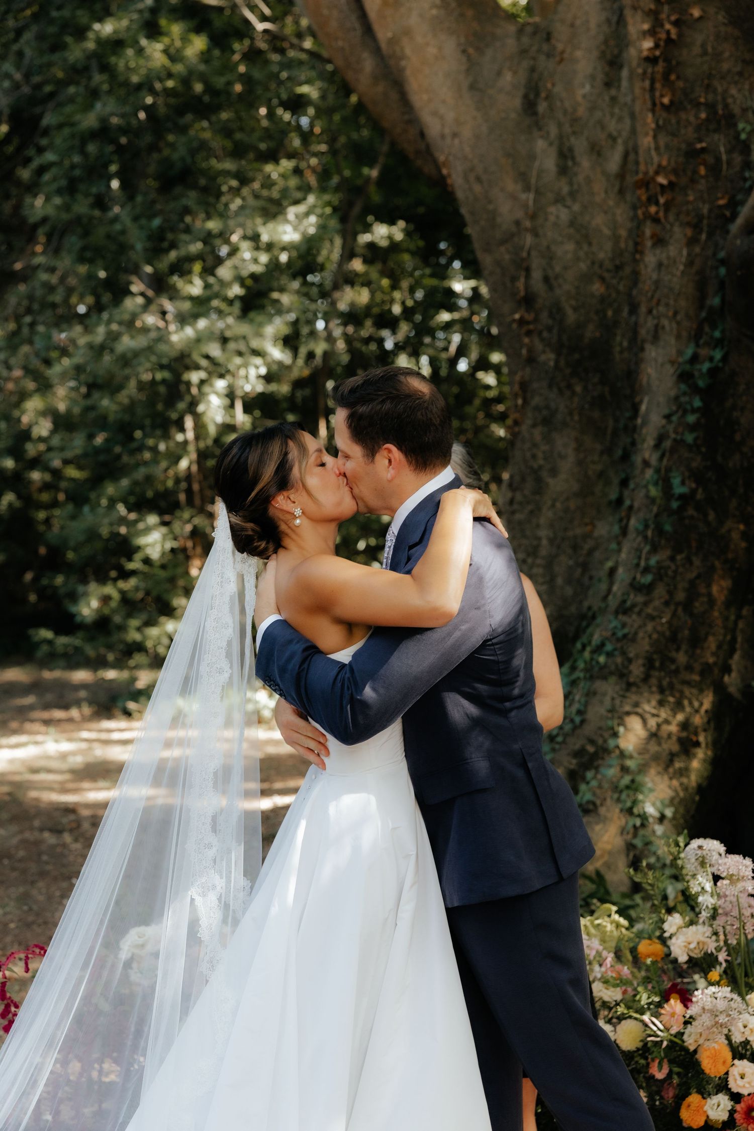 A couple shares their first kiss during an outdoor wedding ceremony surrounded by flowers.