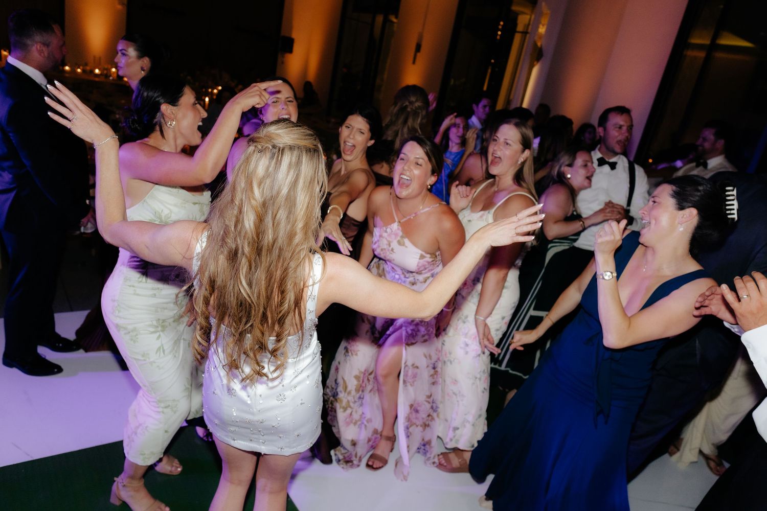 Bride and female wedding guests dance joyfully together on a crowded, colorfully lit reception dance floor.