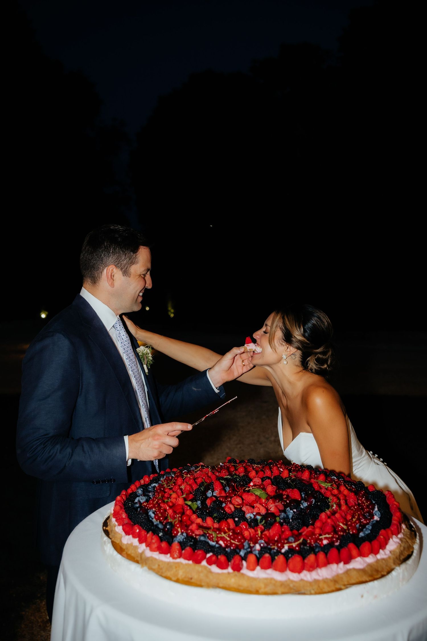 A couple shares a playful moment while cutting into a large berry-topped wedding cake at their reception.