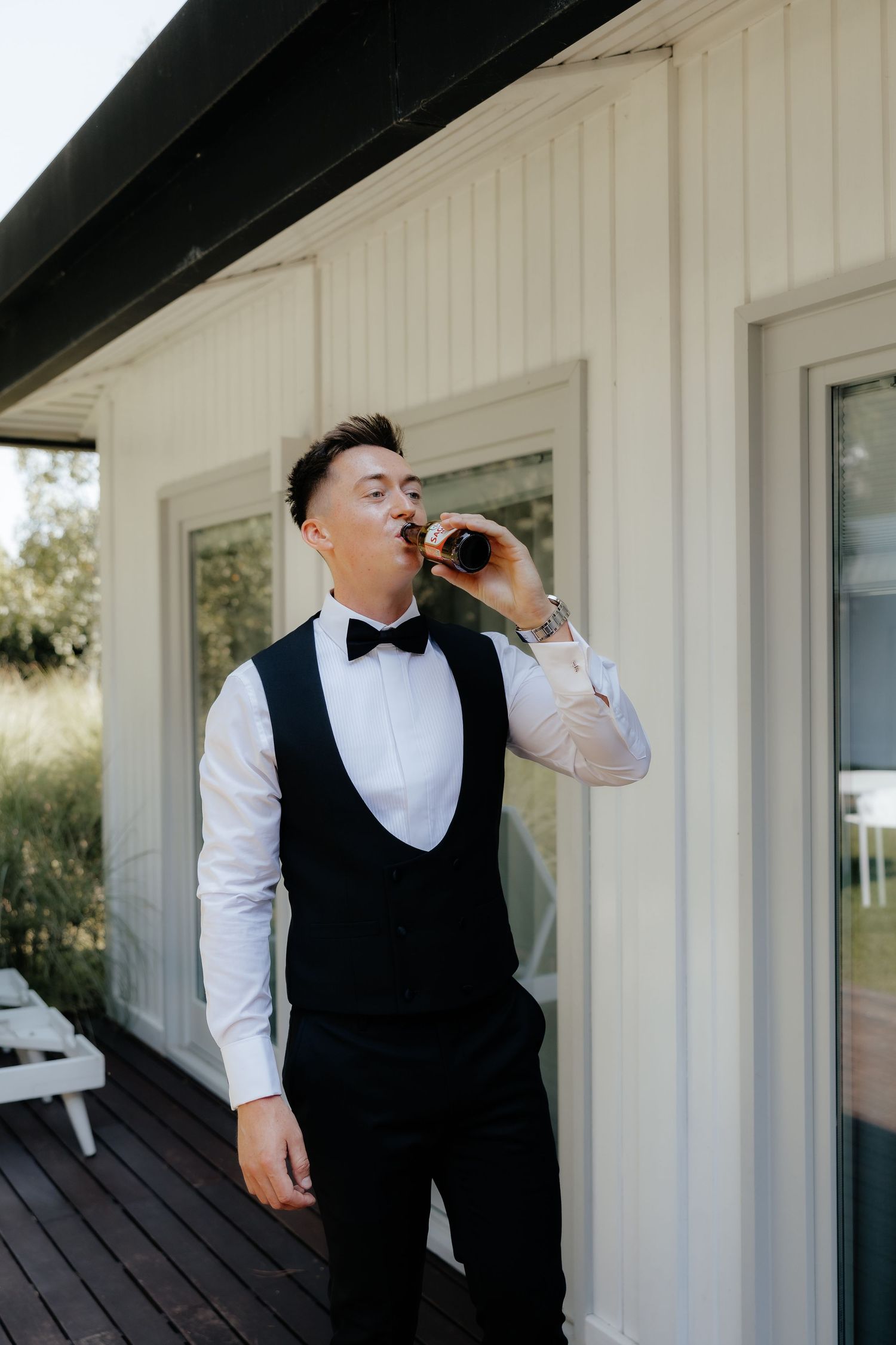 A person in formal attire with a black vest and white shirt stands on a porch and drinks from a glass bottle.