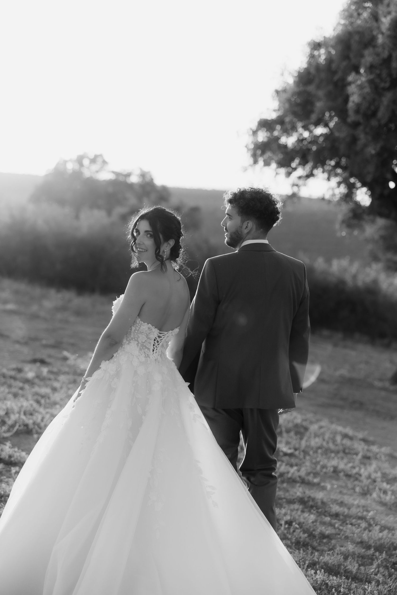 Black and white photo of smiling bride in lace gown walking with groom through sunlit countryside.