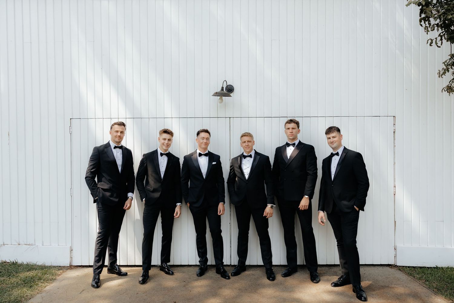 Six groomsmen in black tuxedos posing formally in front of a white barn door.