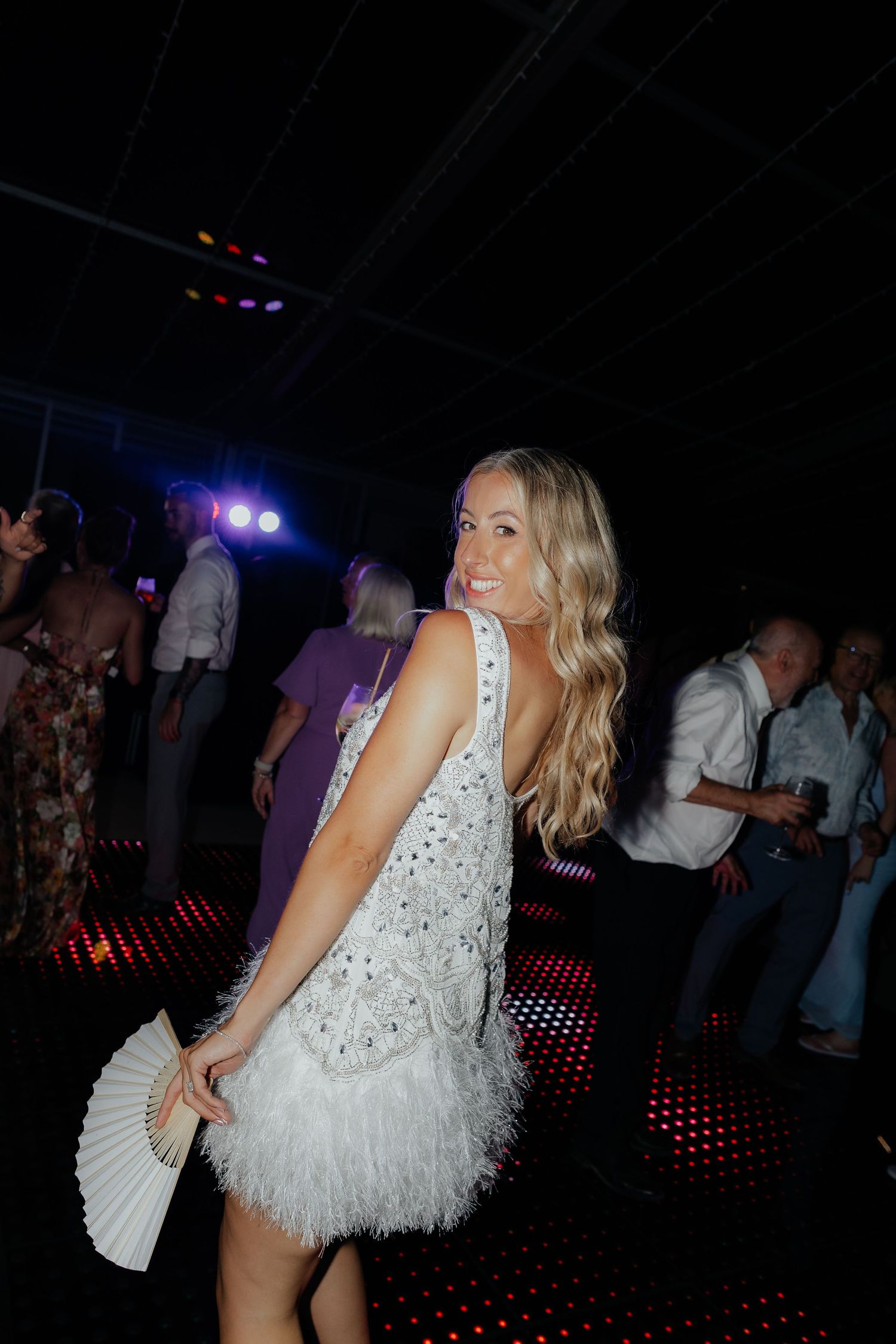 Woman in sparkly white feathered mini dress dancing at a nightclub with a handheld fan.