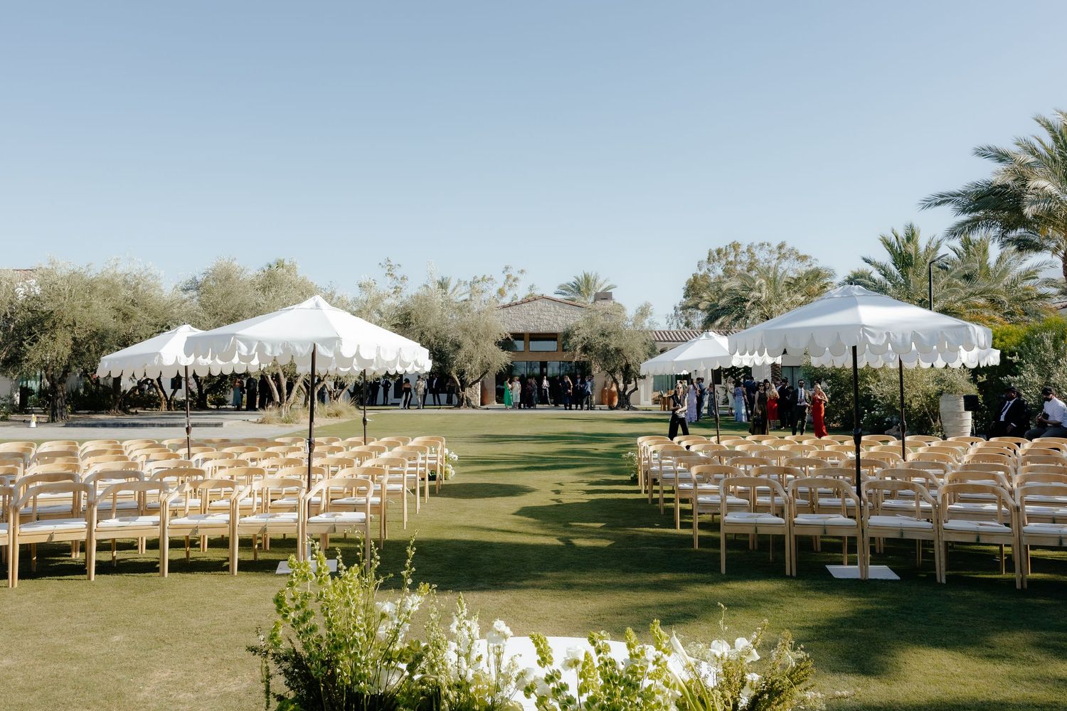 Outdoor wedding ceremony venue with rows of wooden chairs, white scalloped umbrellas, and tall palm trees under blue sky.