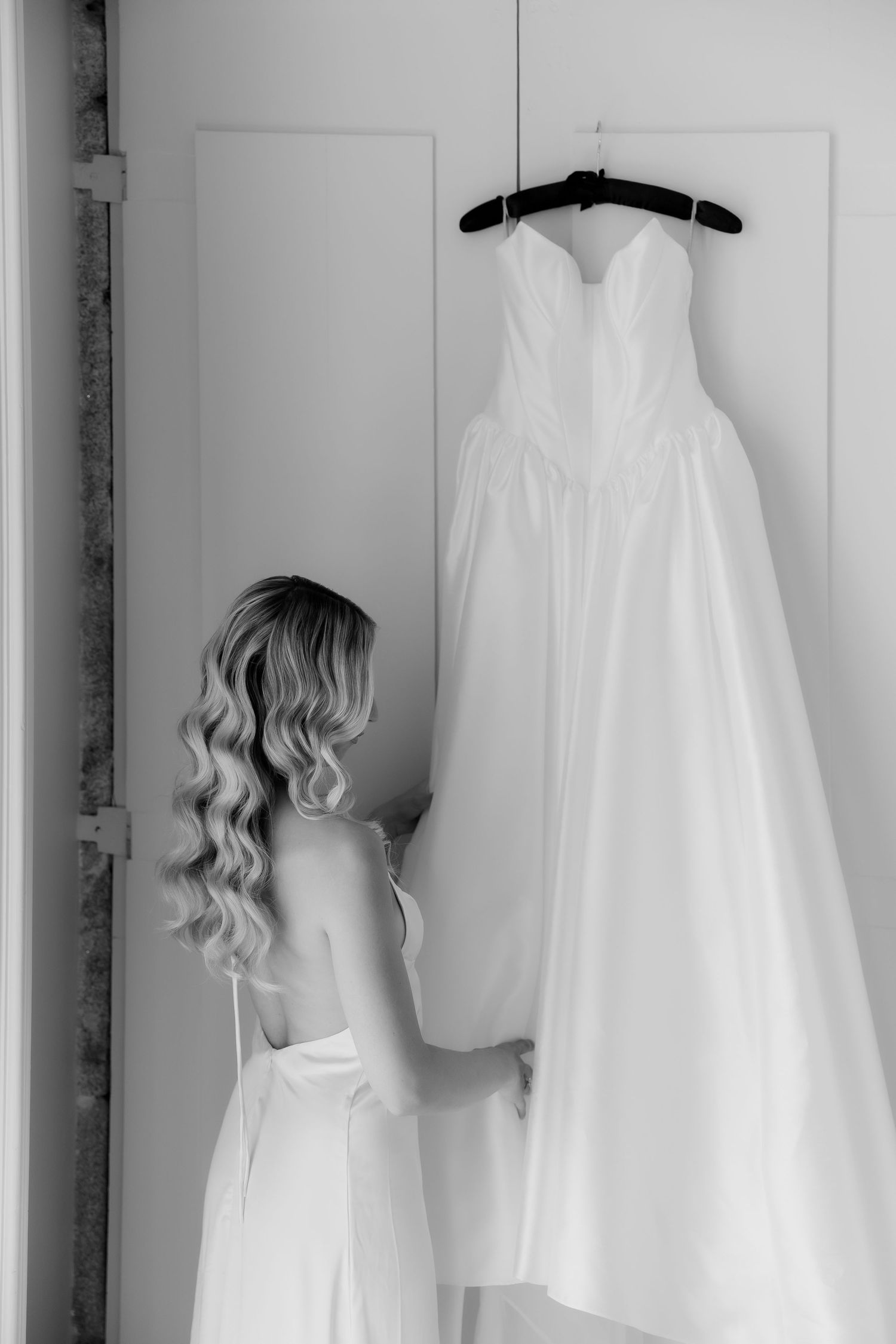 Black and white rear view of bride in slip dress reaching for hanging wedding gown.
