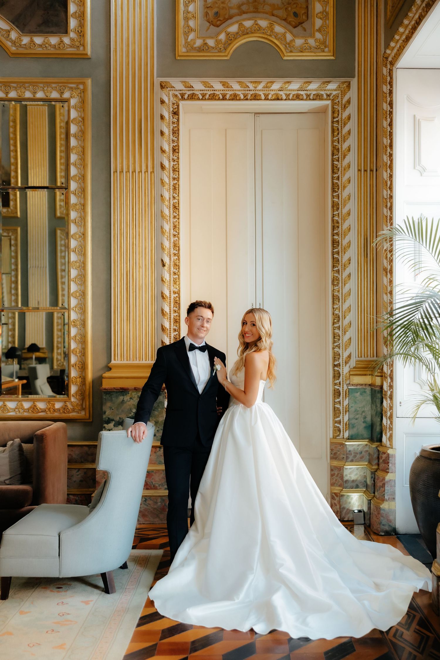 Bride and groom posing in an opulent gold-adorned ballroom, bride wearing a flowing white ball gown.