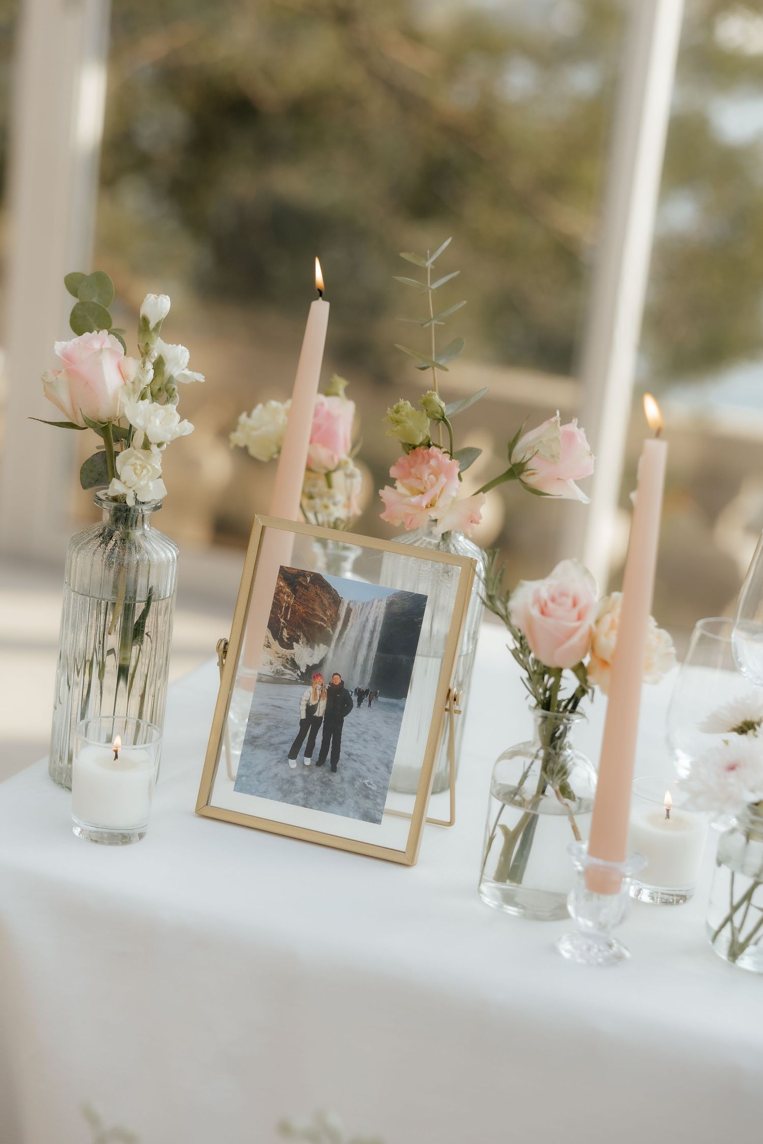Wedding reception table decorated with pink roses, framed photos, and white candles in an elegant table setting.
