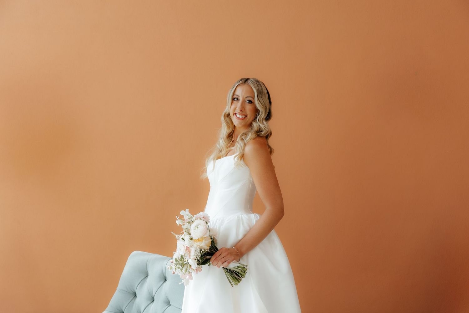 A bride in a white strapless wedding gown holds a bouquet while standing beside a light blue tufted chair.