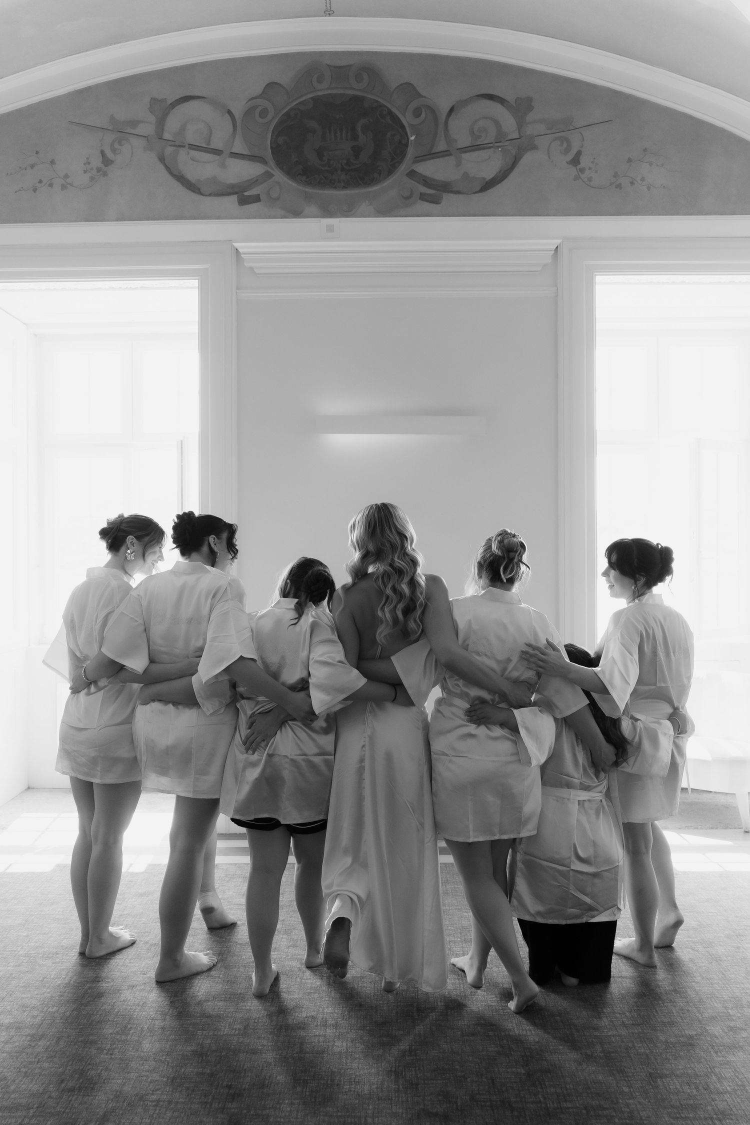 A group of dancers in white outfits lean against a wall in sequence, creating an artistic formation in a bright studio space.