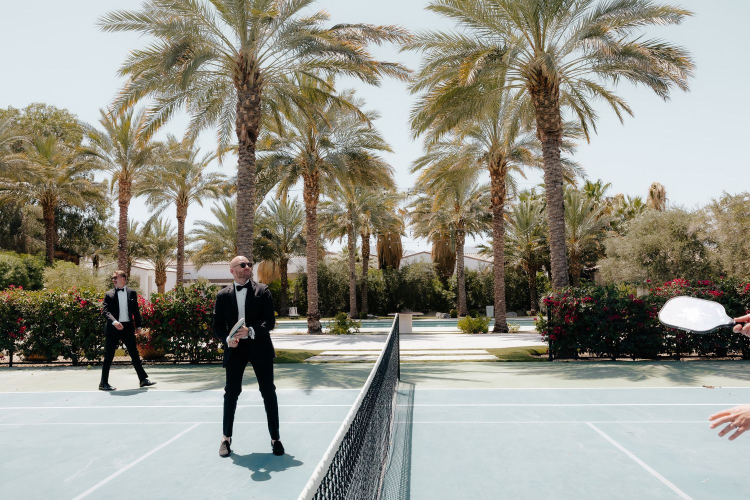 Groom playing tennis in black tuxedo on outdoor court surrounded by palm trees.