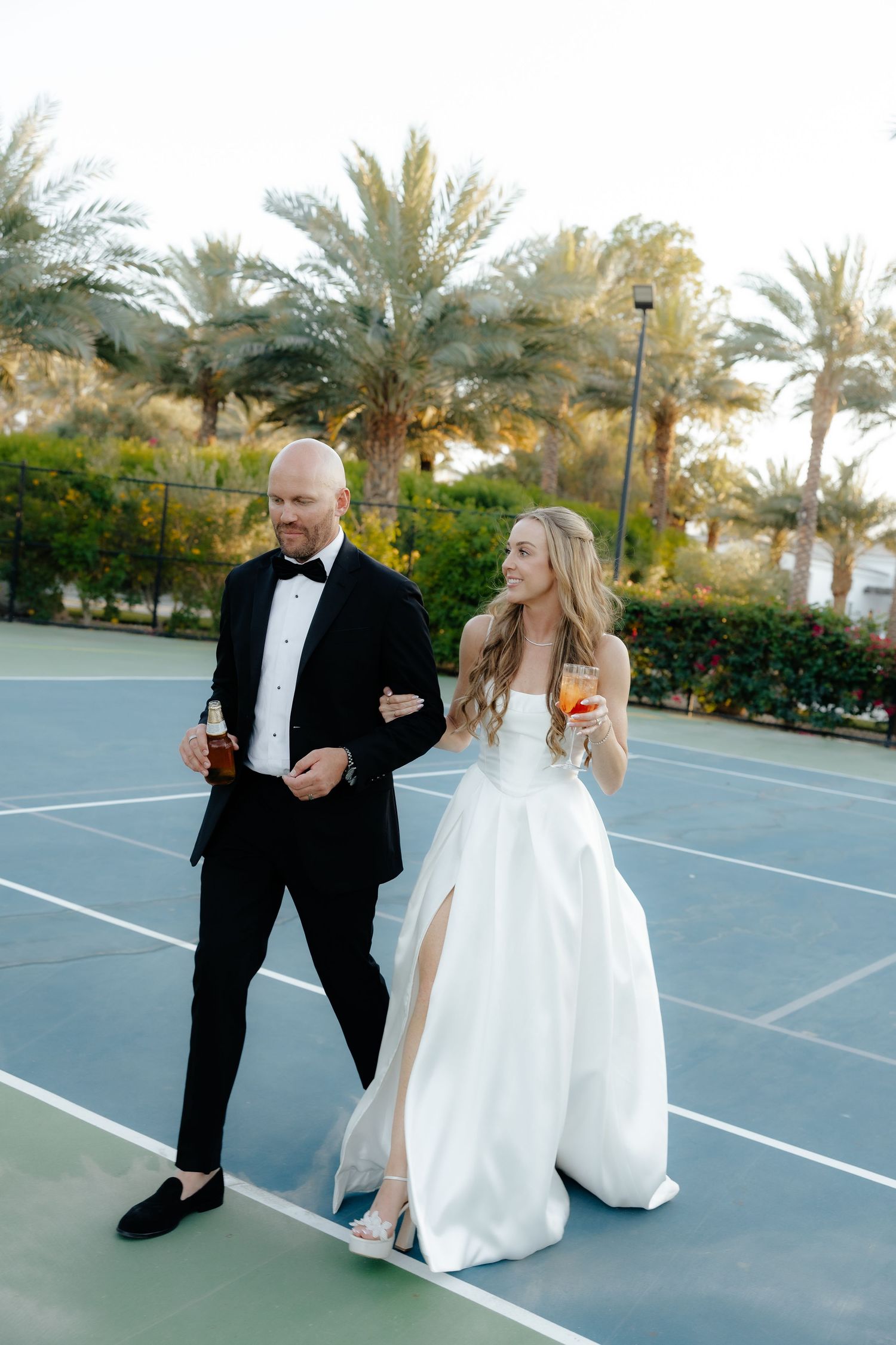 Bride and groom walking on tennis court holding cocktails, surrounded by palm trees at outdoor wedding venue.