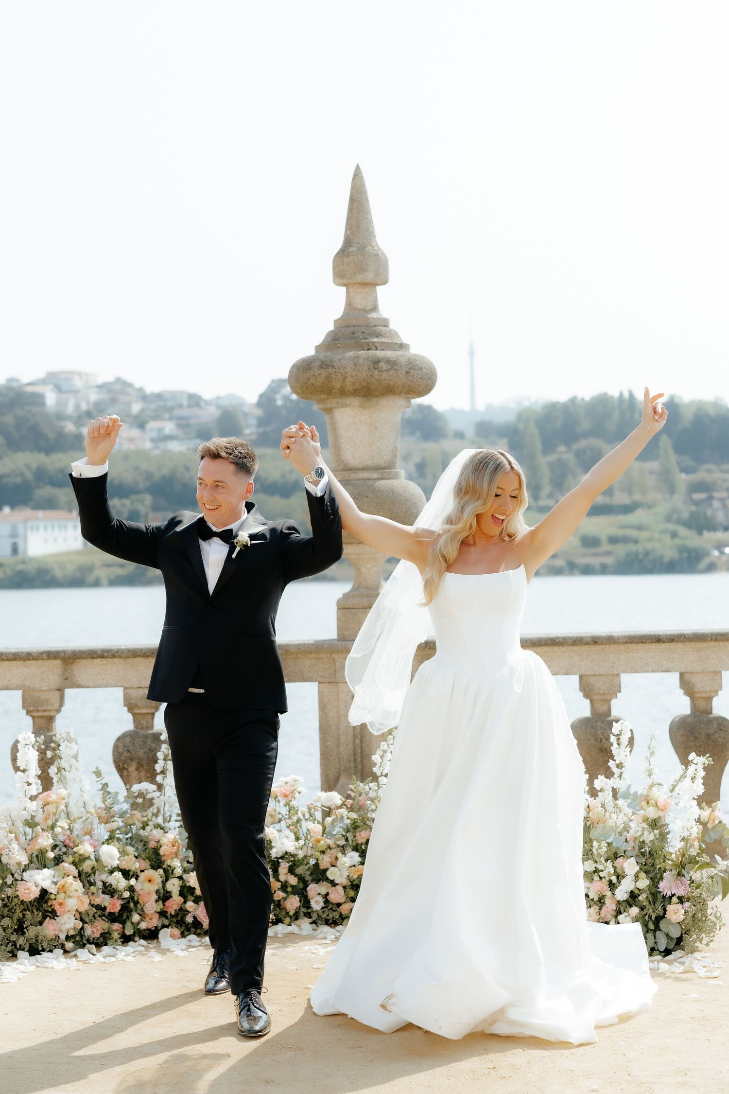 A joyful couple in wedding attire celebrates with raised arms on a scenic balcony overlooking water and gardens.