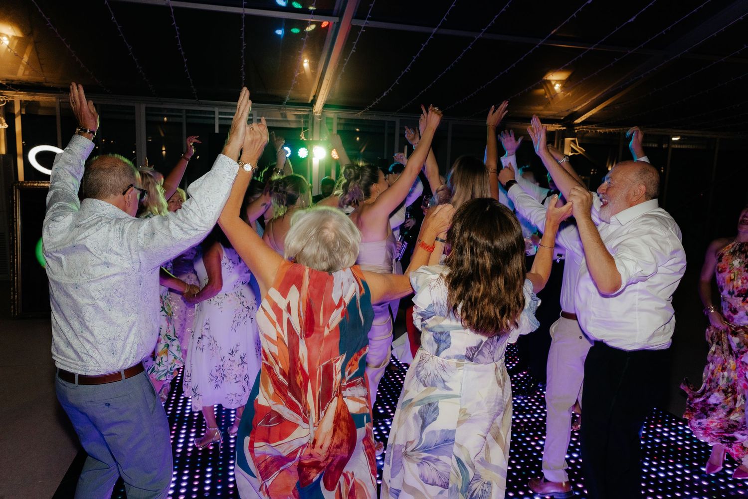 Party guests dance with hands raised on an illuminated dance floor under colorful purple lighting at a celebration event.