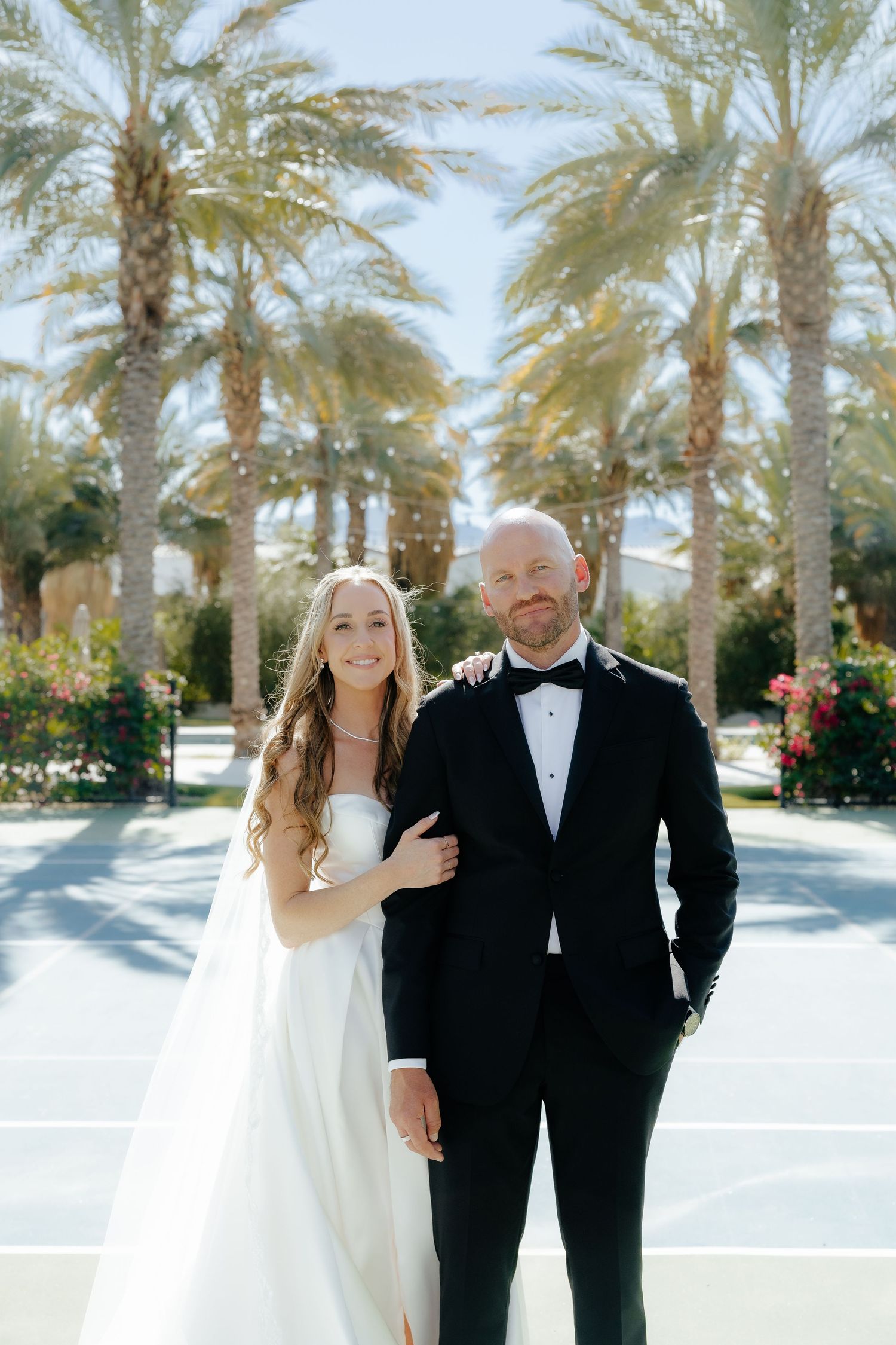 Bride and groom pose together in front of tall palm trees, she in a white strapless gown, he in a black tuxedo.