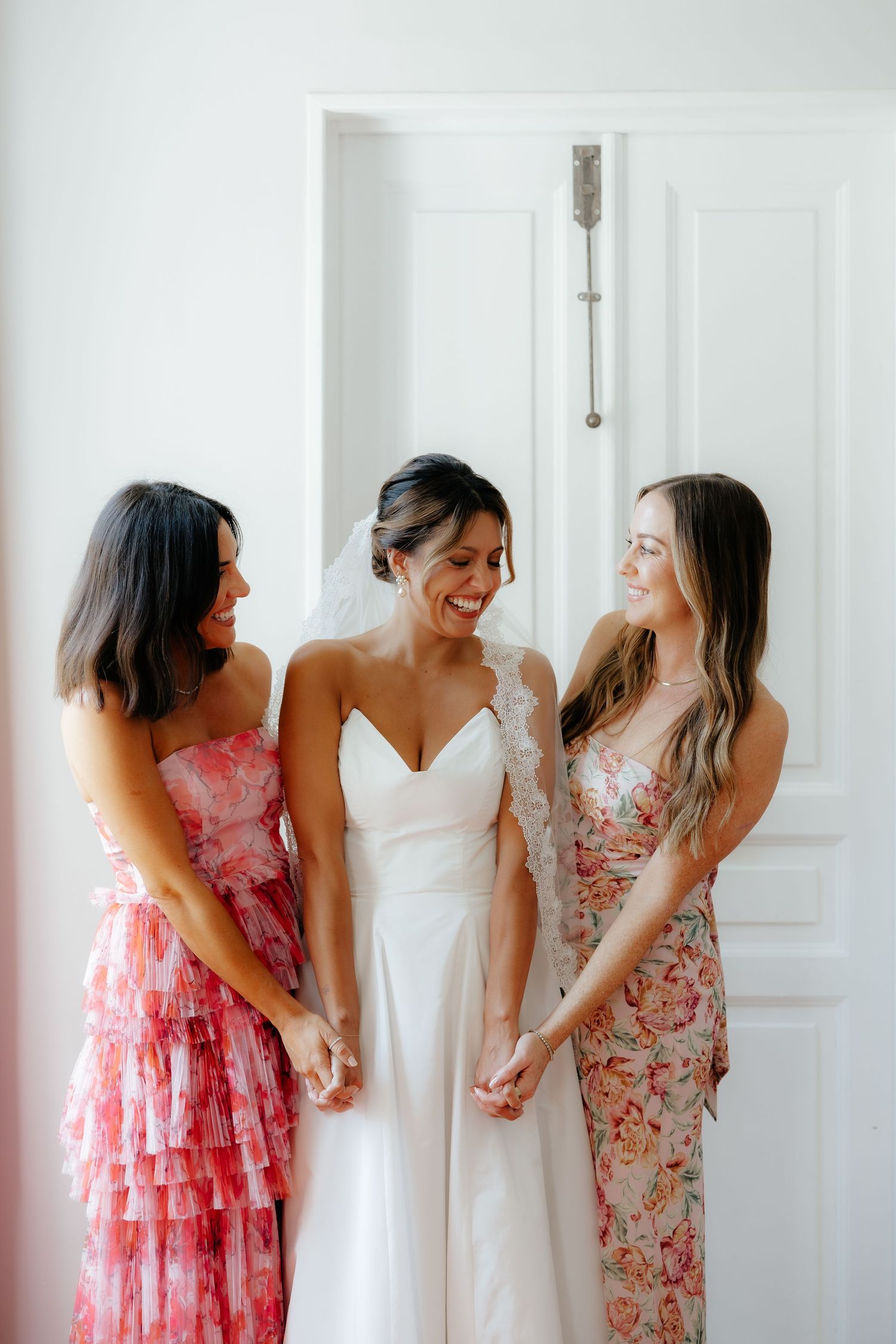 Three women in formal dresses share a joyful moment together against a white door background.