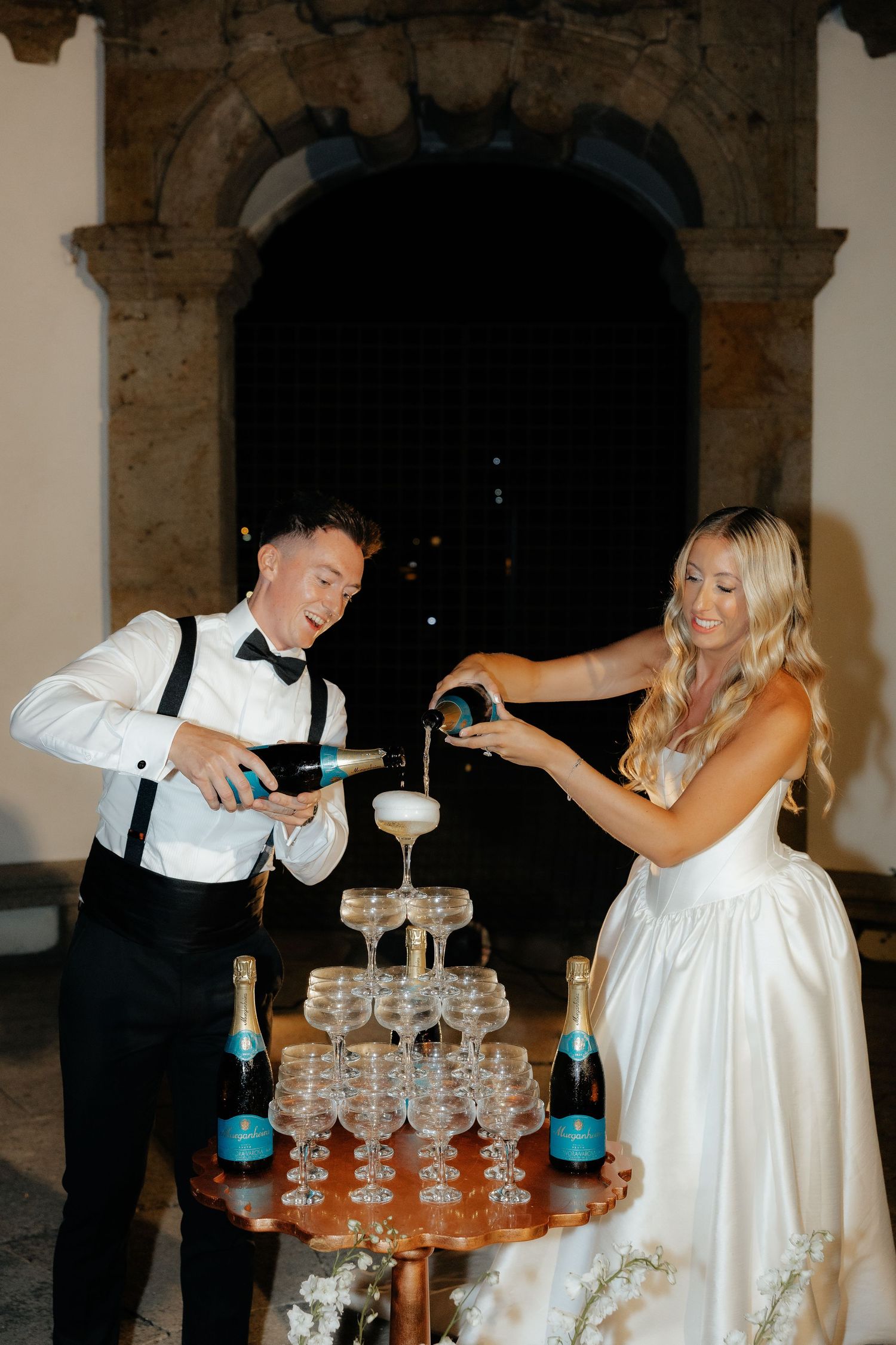 A couple pours champagne into a tower of coupe glasses at an elegant wedding reception.