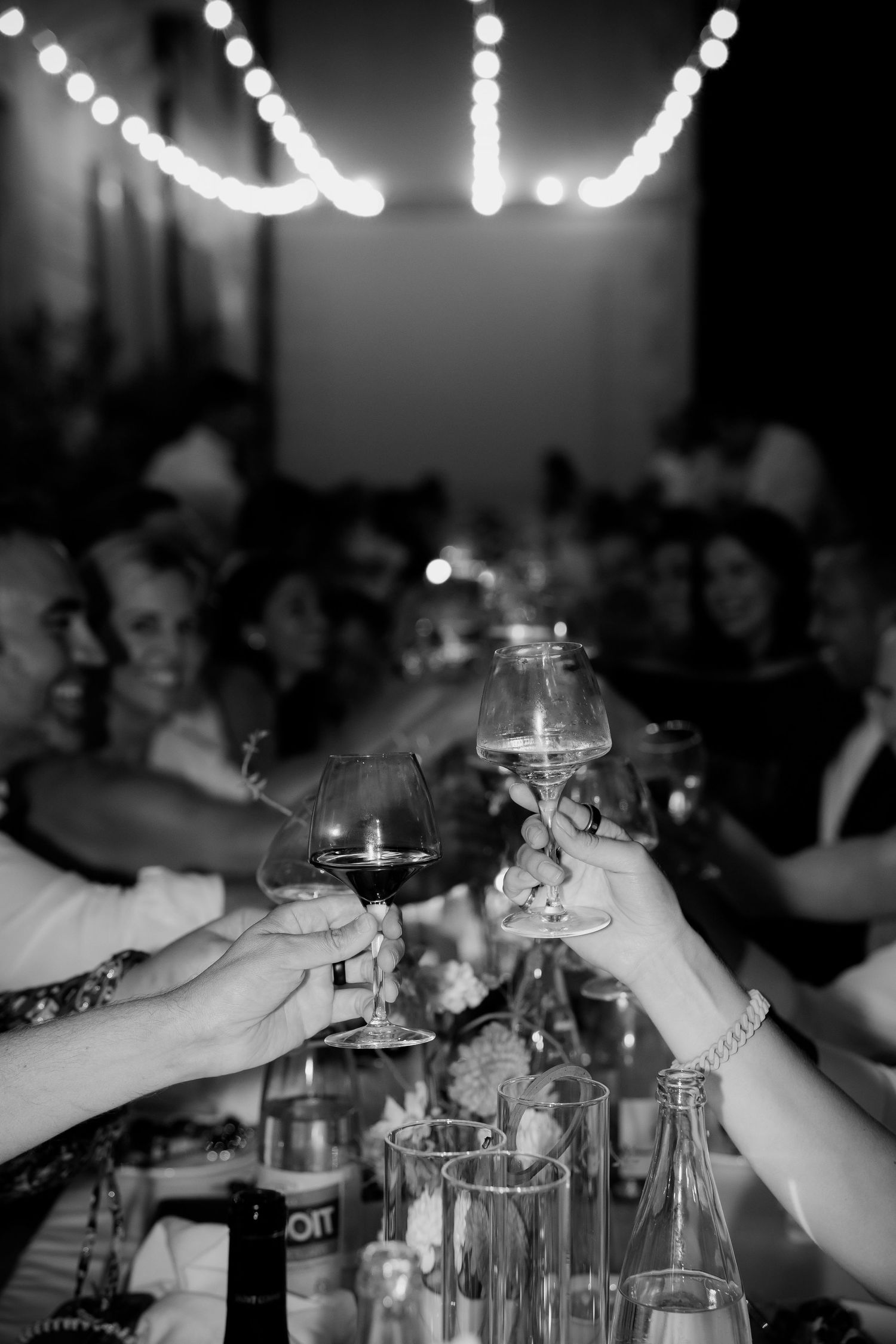 Wedding guests raise their glasses in a celebratory toast during an evening reception with bokeh lights in background.