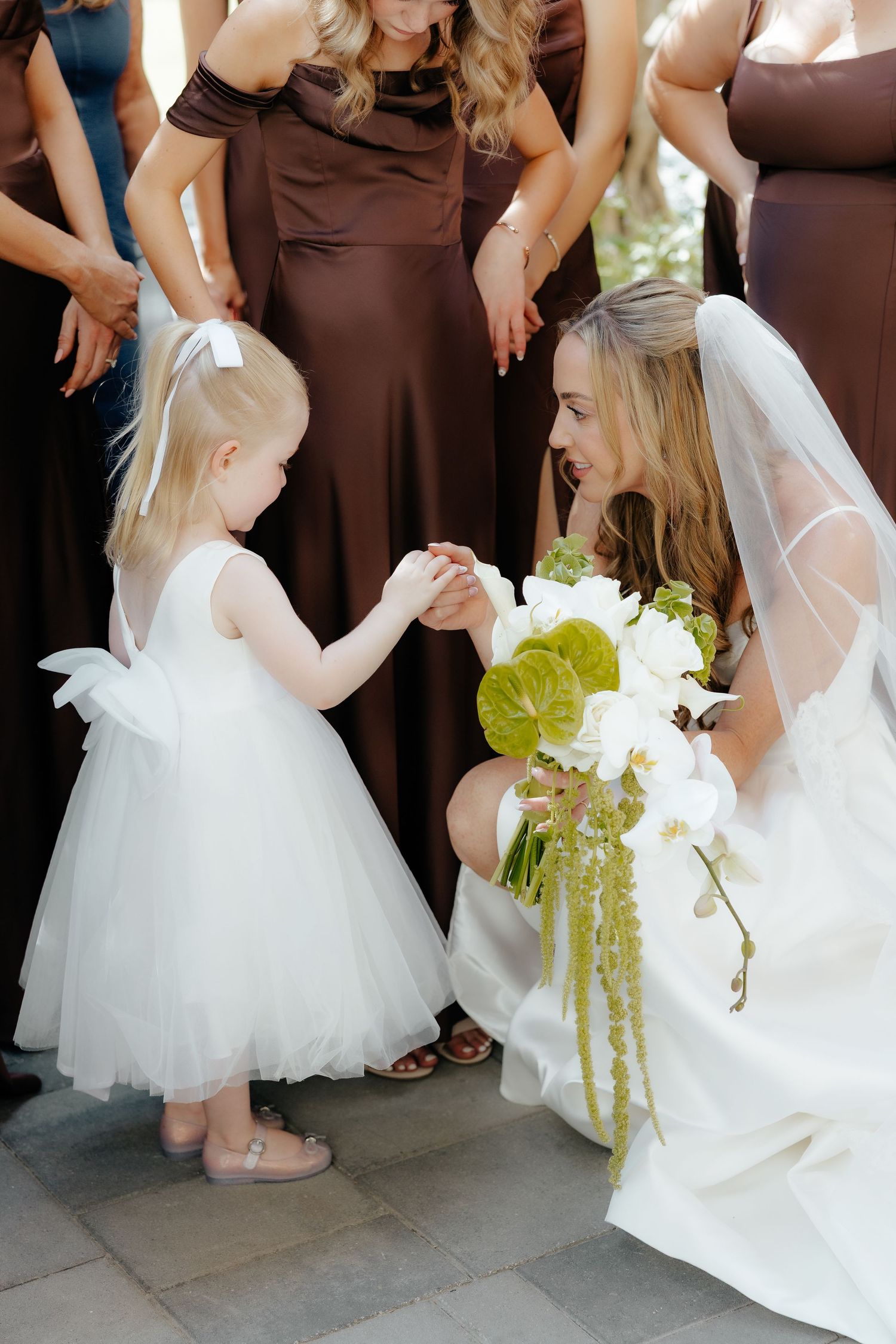 Bride in white wedding dress holding bouquet while interacting with a flower girl outdoors.