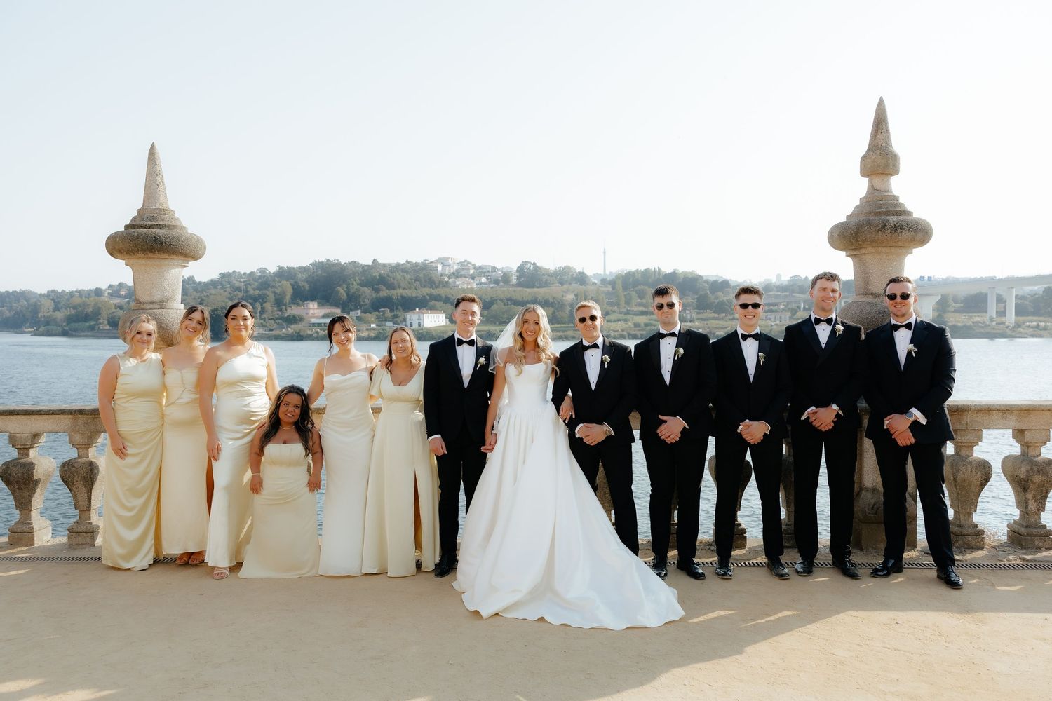 A wedding party poses on an elegant waterfront terrace with ornate stone balustrades and a scenic bay view.