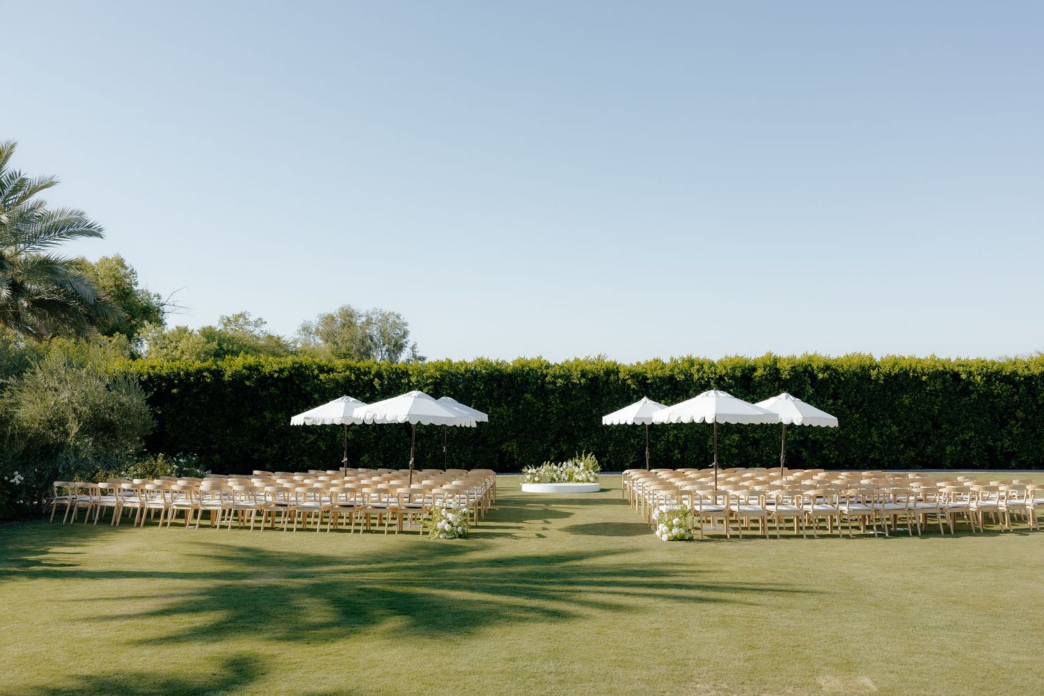 Outdoor wedding ceremony setup with rows of chairs and white umbrellas on green lawn.
