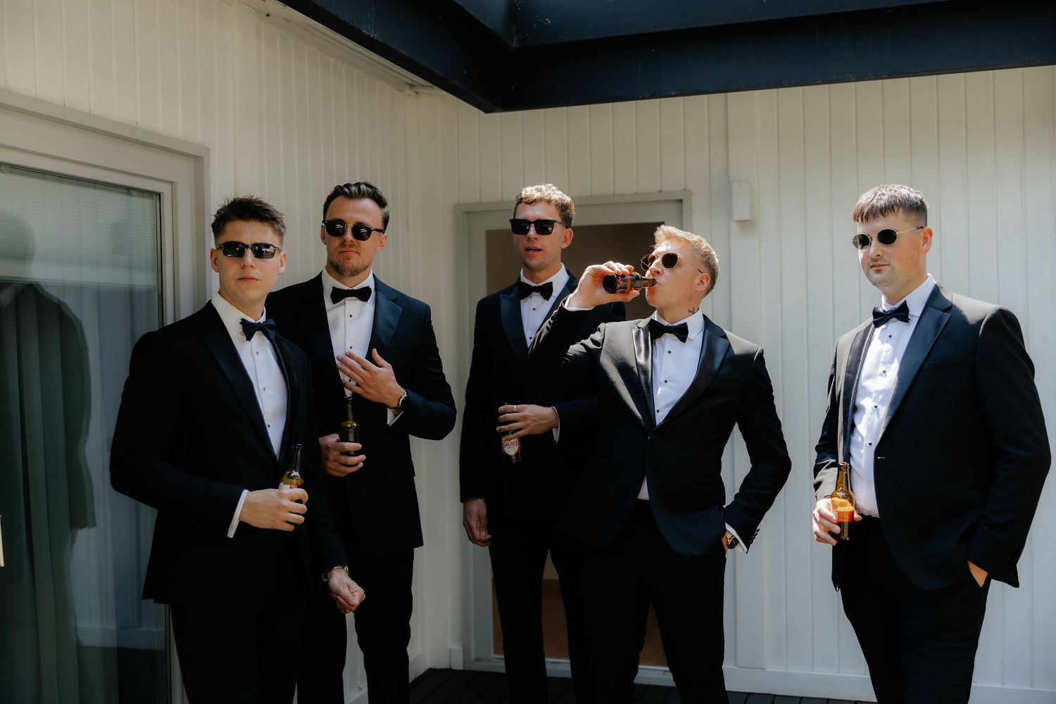 A group of groomsmen in black tuxedos and sunglasses pose together before a wedding ceremony.