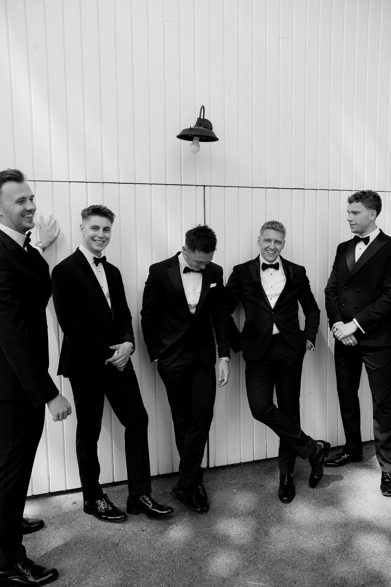 Five groomsmen in black tuxedos laughing and posing against a white barn wall.