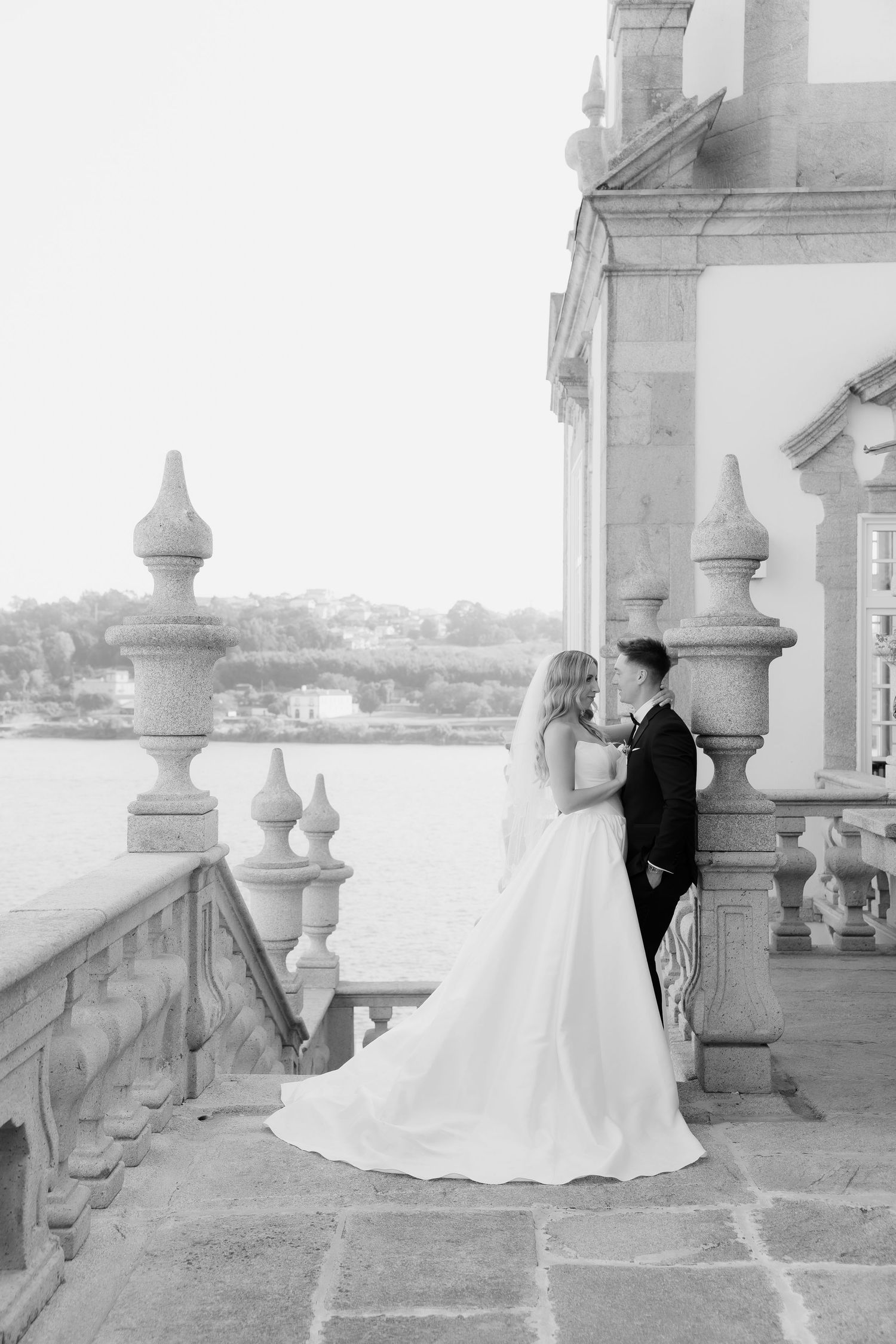 Bride and groom share romantic moment on ornate stone balcony with waterfront view in black and white.