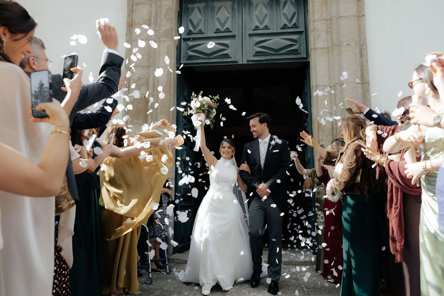Bride and groom exit church doors as guests throw white confetti petals in joyful wedding celebration.