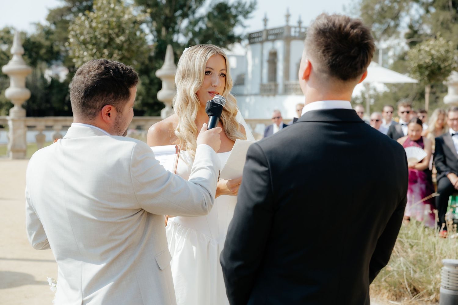 A wedding celebration takes place outdoors on a sunny day with elegant architecture in the background.