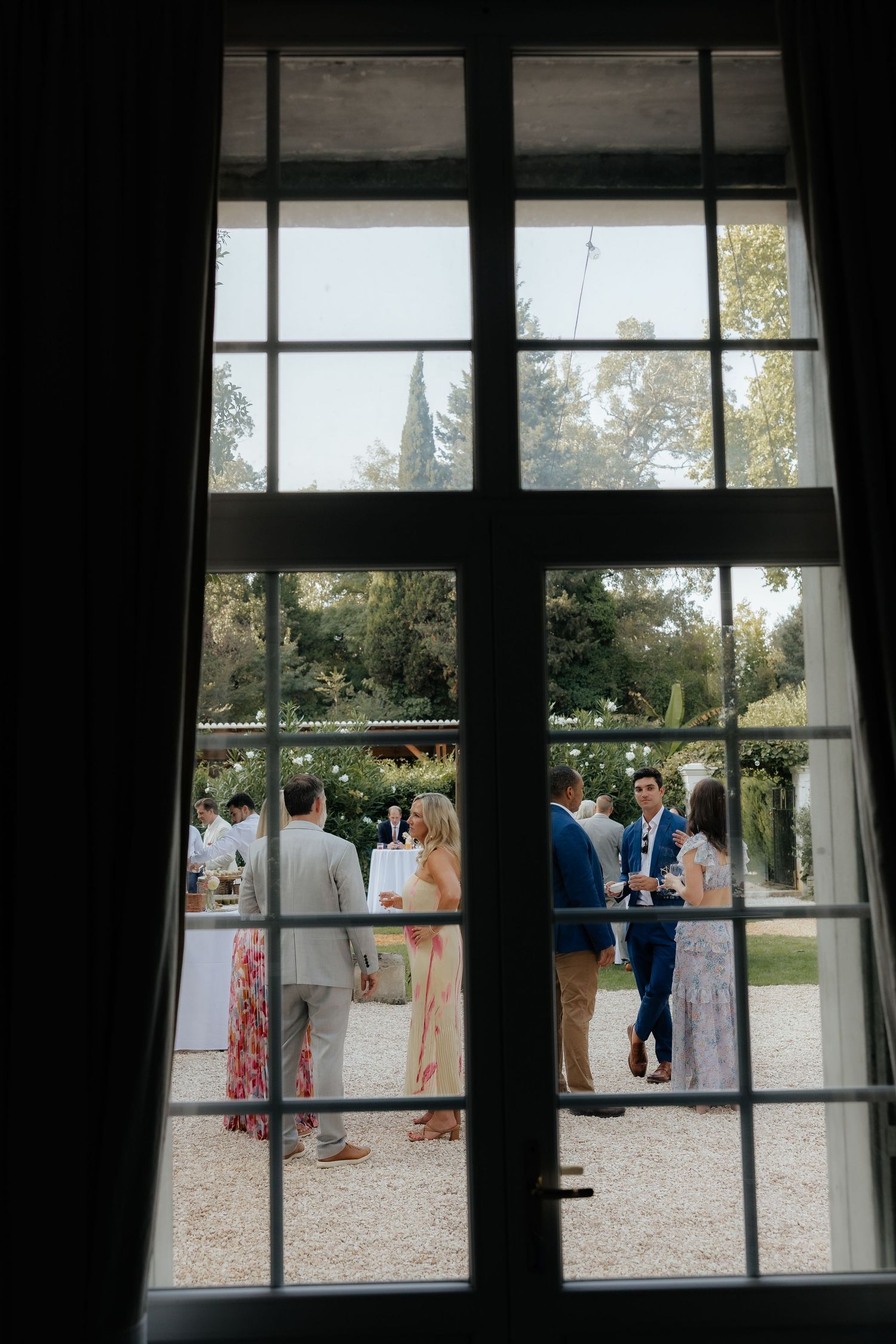 View through dark window panes of wedding guests mingling on a sunny outdoor patio.