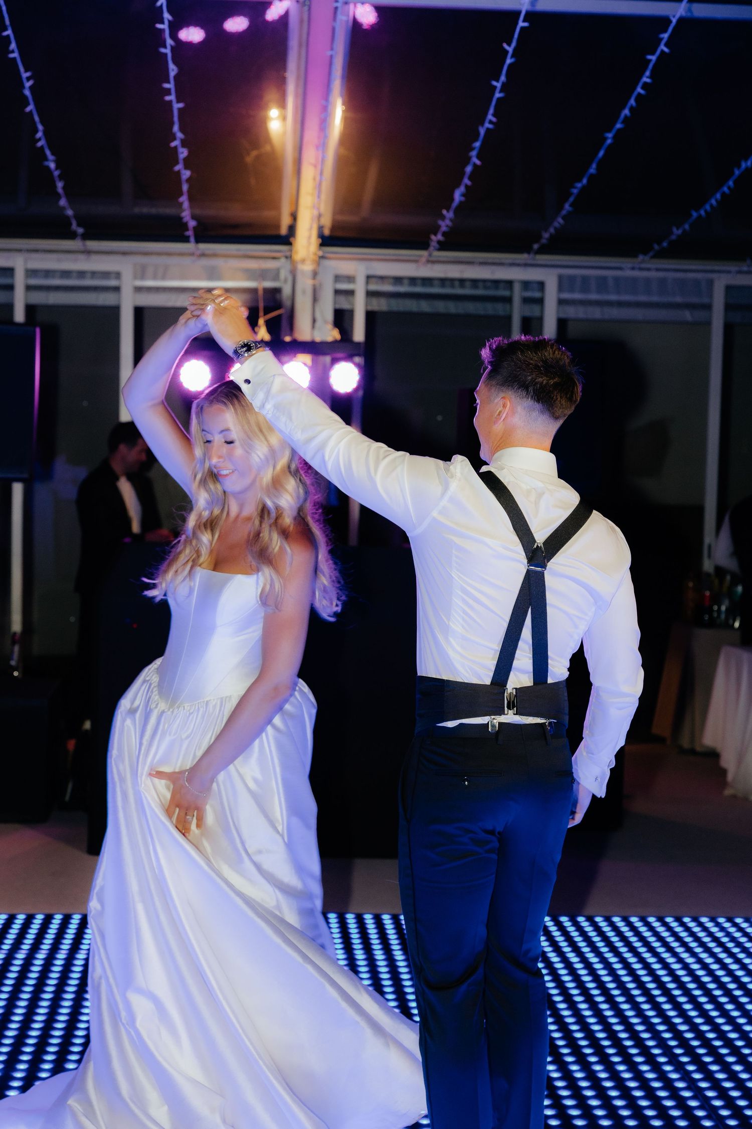 Newlywed couple dancing on illuminated checkerboard dance floor under purple lighting at wedding reception.