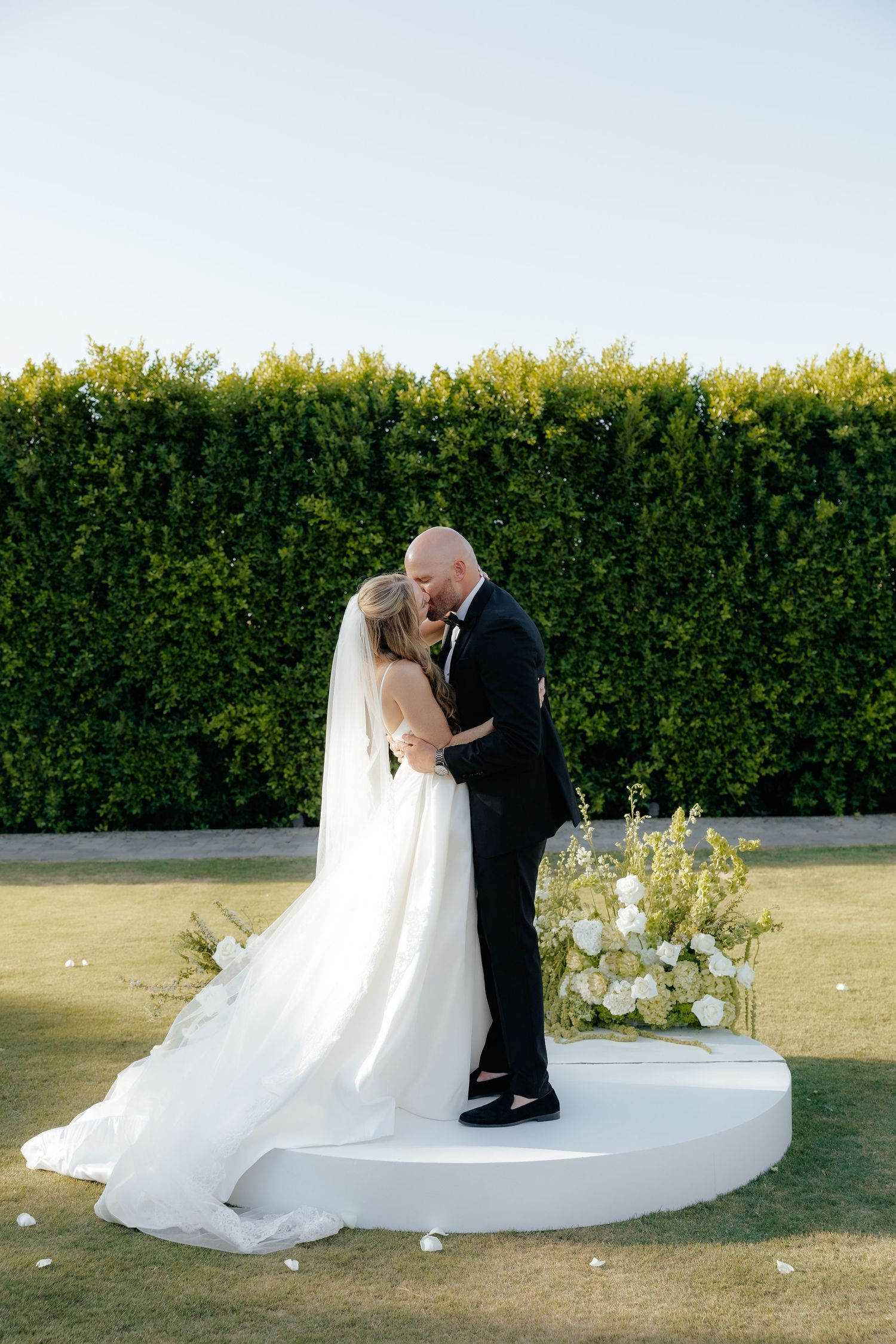 Bride and groom share first kiss on white circular platform with white floral arrangements and green hedge backdrop.