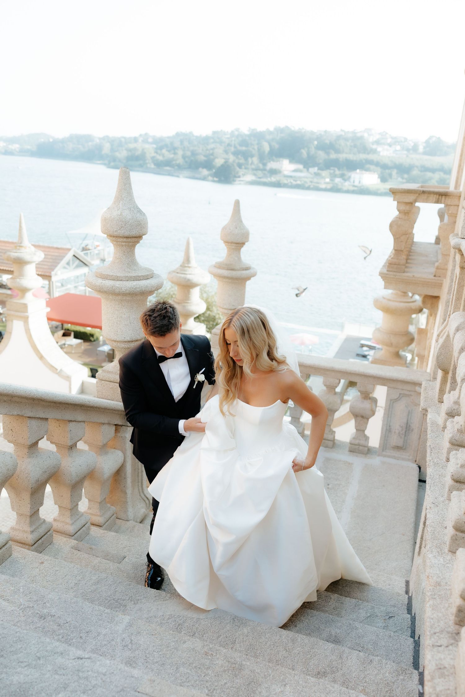 Newlywed couple in formal attire sits on ornate stone balustrade overlooking scenic coastal view.
