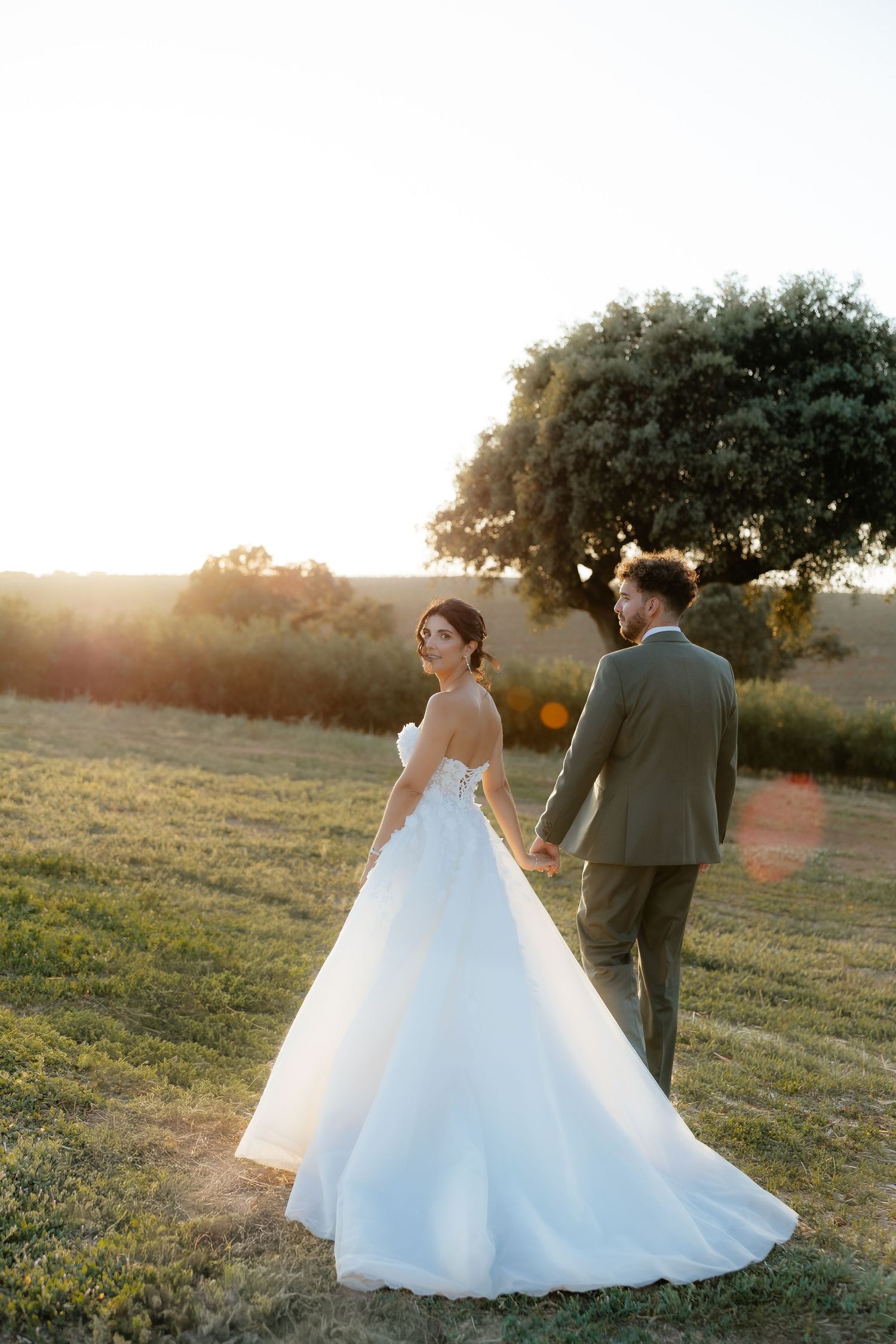 Bride in flowing white ballgown and groom in olive suit holding hands at golden hour in open field.