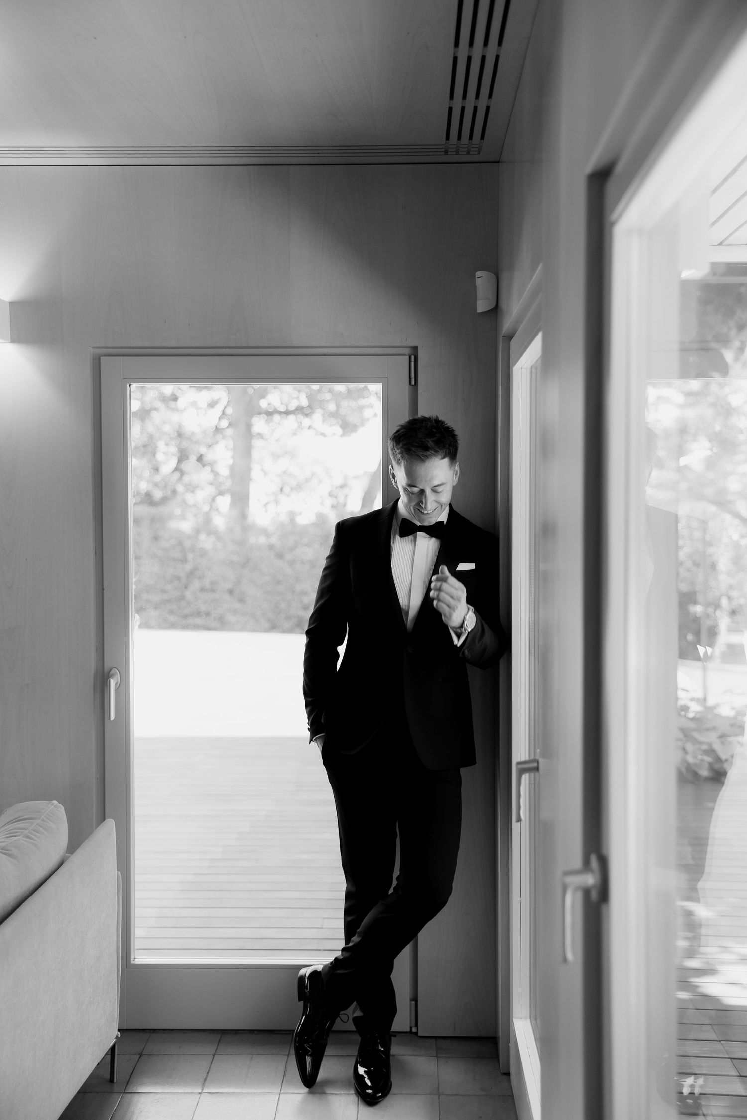 Black and white portrait of a groom in a tuxedo leaning against a door frame adjusting his jacket indoors.