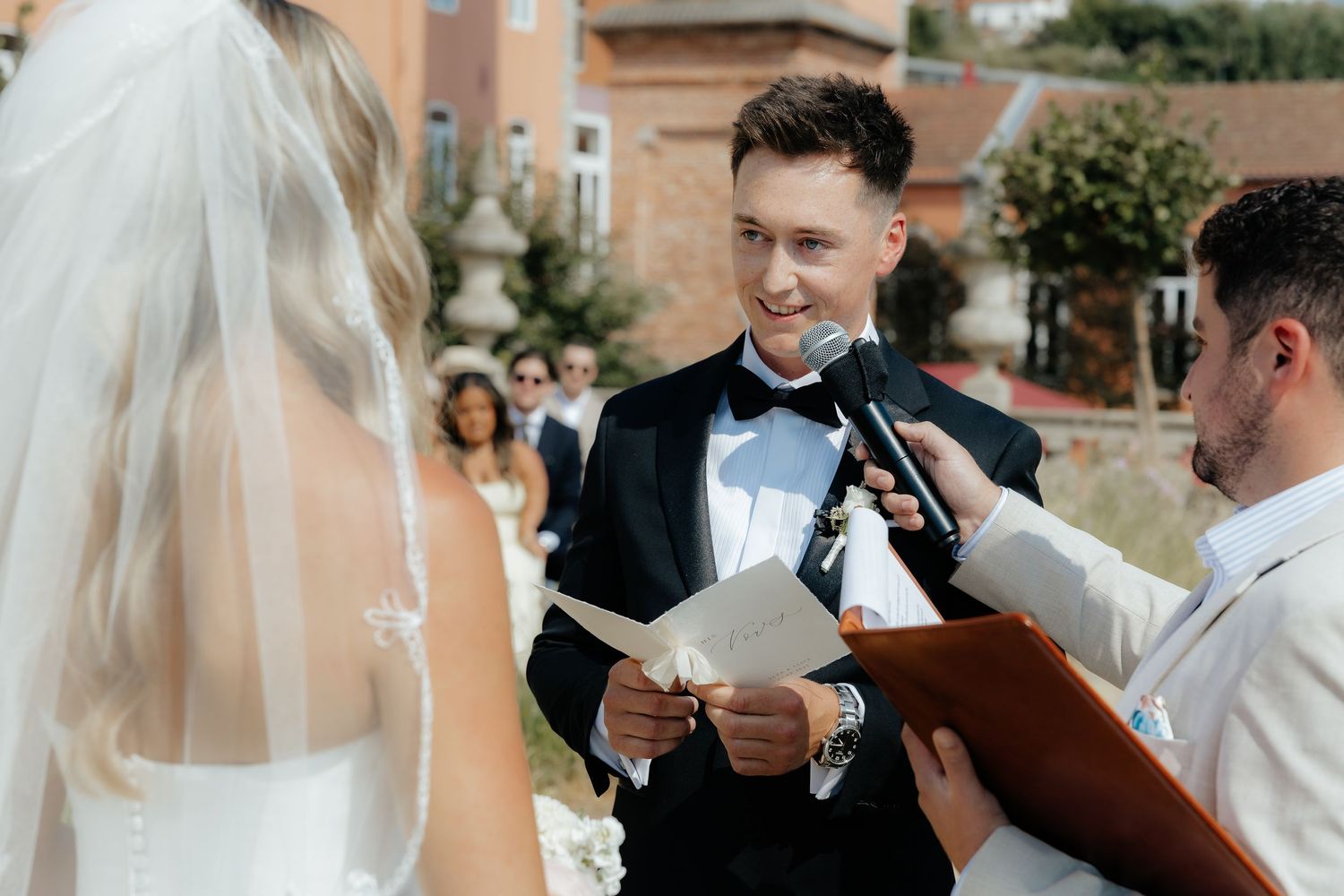 Groom smiles reading heartfelt vows to bride at elegant outdoor wedding ceremony.