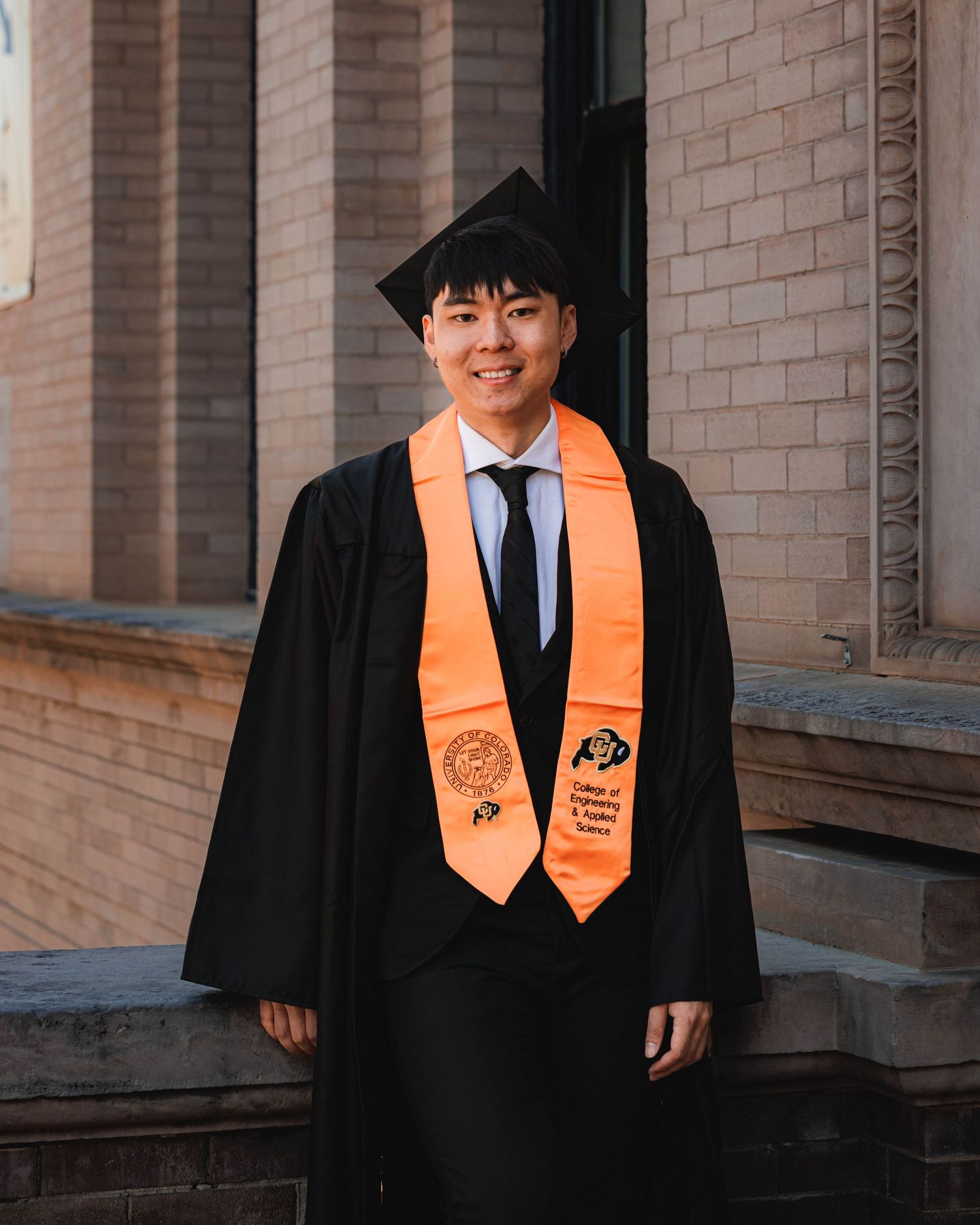 CU Boulder graduate in cap and gown posing on campus near a campus building