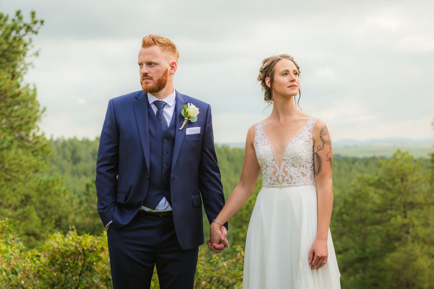 Wolfe Wedding in Franktown, Colorado, newly weds holding hands, looking off in opposite directions, with Colorado mountains and trees behind