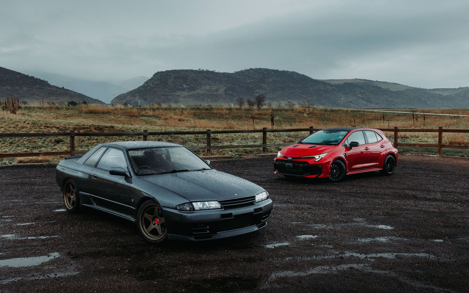 Wide angle shot of Gunmetal Grey 1990 Nissan Skyline GTR R32 next to Red 2024 GR Corolla with mountains in background during a rain storm