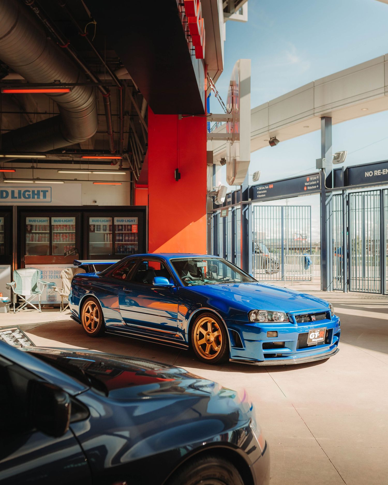 Blue Nissan Skyline GTR R34 on gold TE37s, at Down To Earth Movement, inside of Sports Authority Stadium, during sunset.