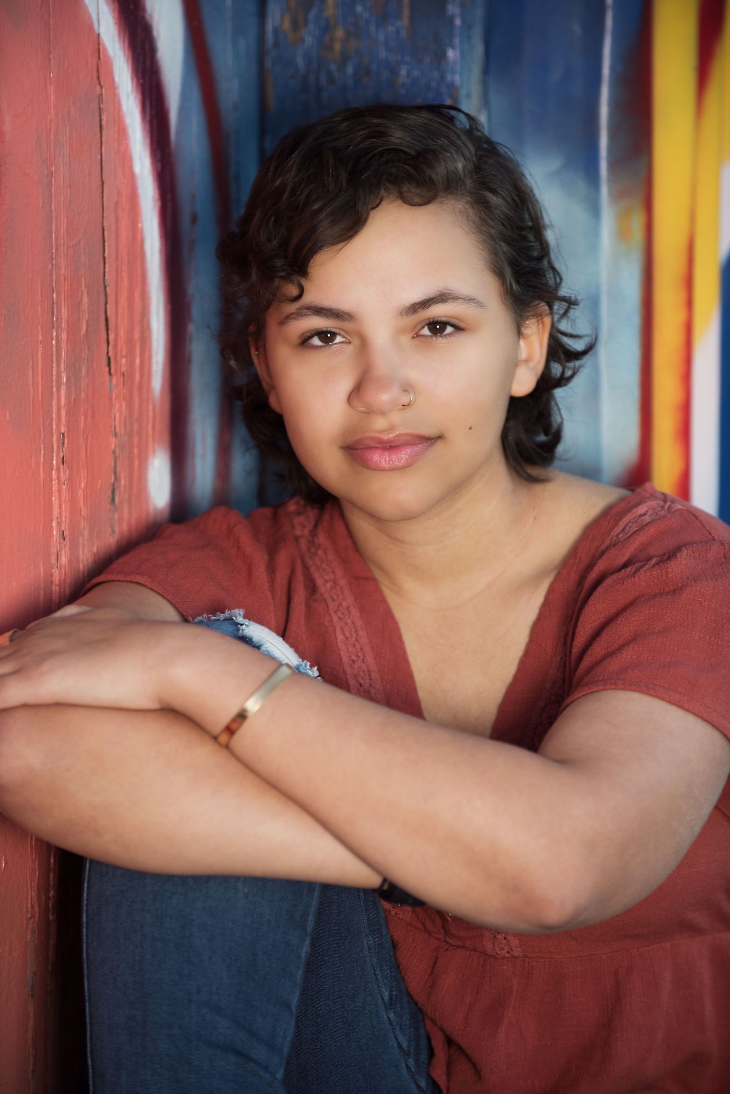 Colorado Springs senior portrait of a student seated beside a colorful rustic wall with natural expression and eye contact.