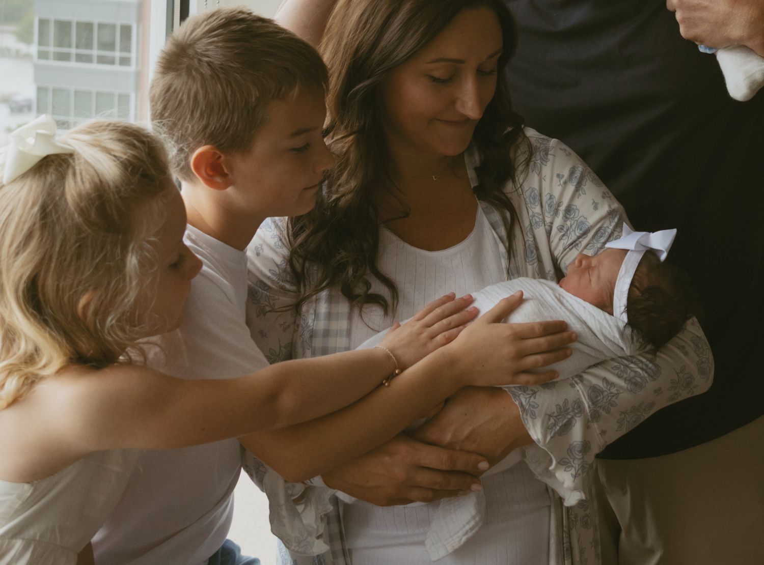 Hudson Valley newborn indoor photo next to window. Kids reaching for newborn baby as mom holds baby in her arms