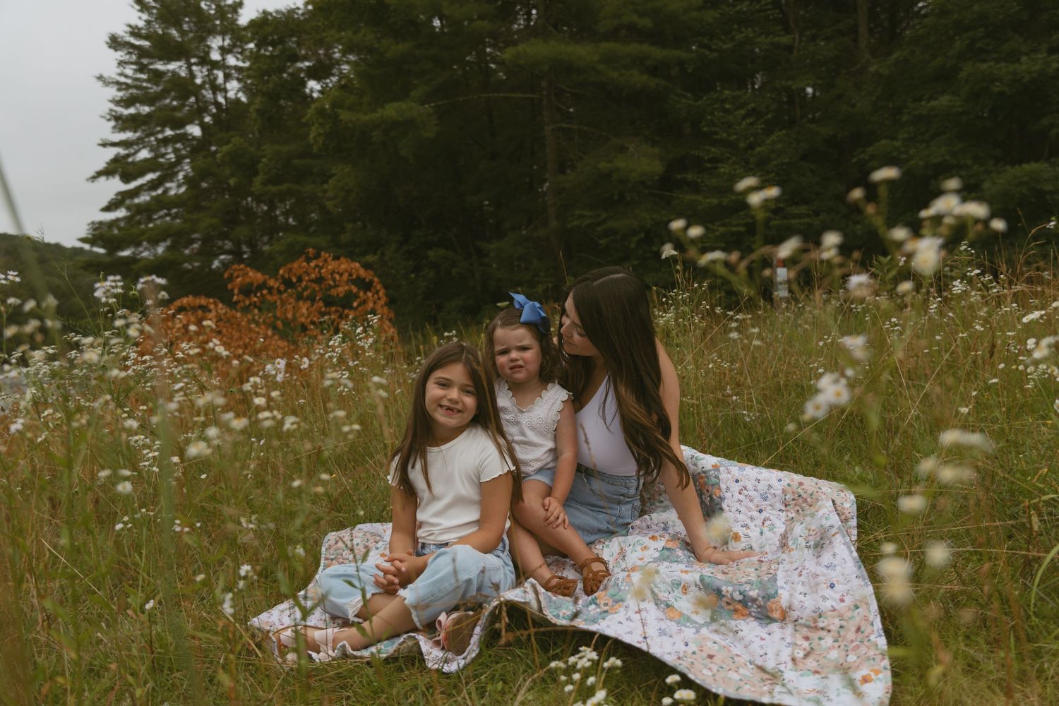 A motherhood photo of a mom sitting in a flower field with her two daughters on a picnic blanket in Orange County New Jersey.