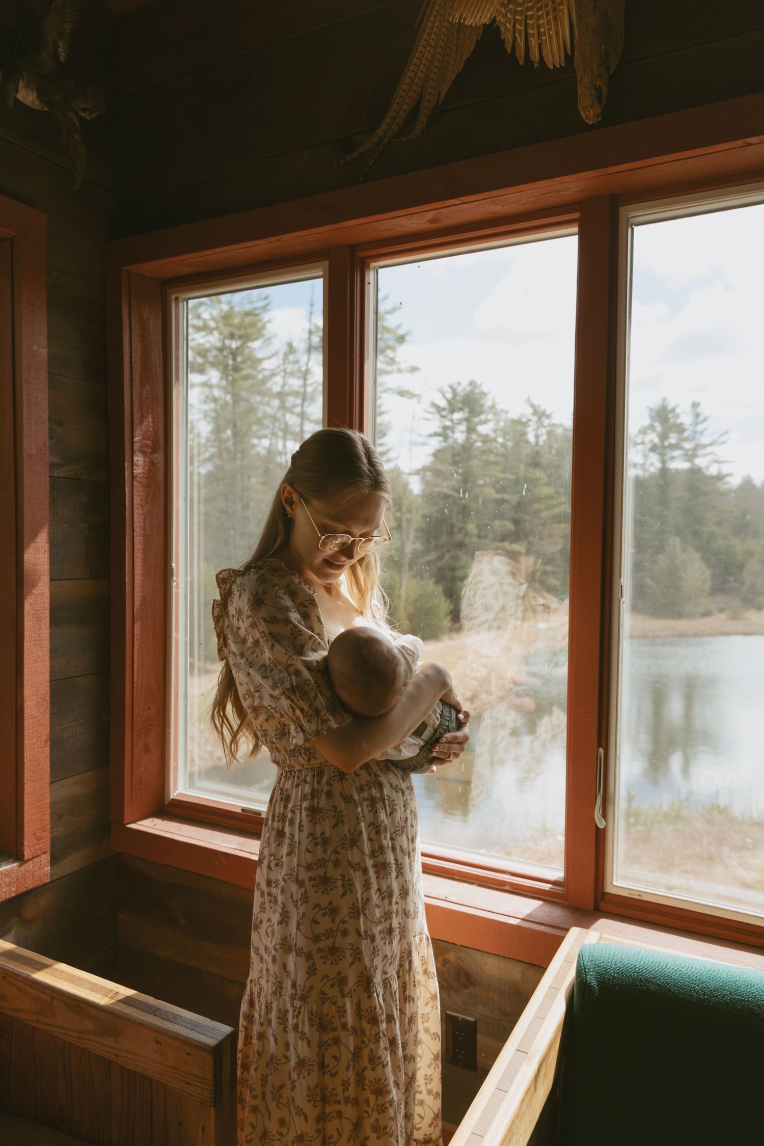 Newborn photos in family cabin in Sullivan County New York, Catskills. Mom next to window as she holds newborn baby.
