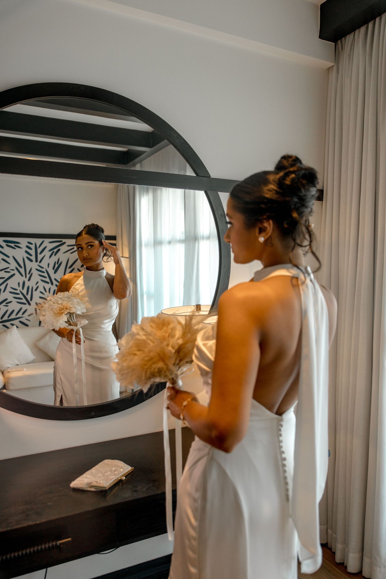 Bride in backless white halter gown holds pampas bouquet while viewing reflection in round mirror.