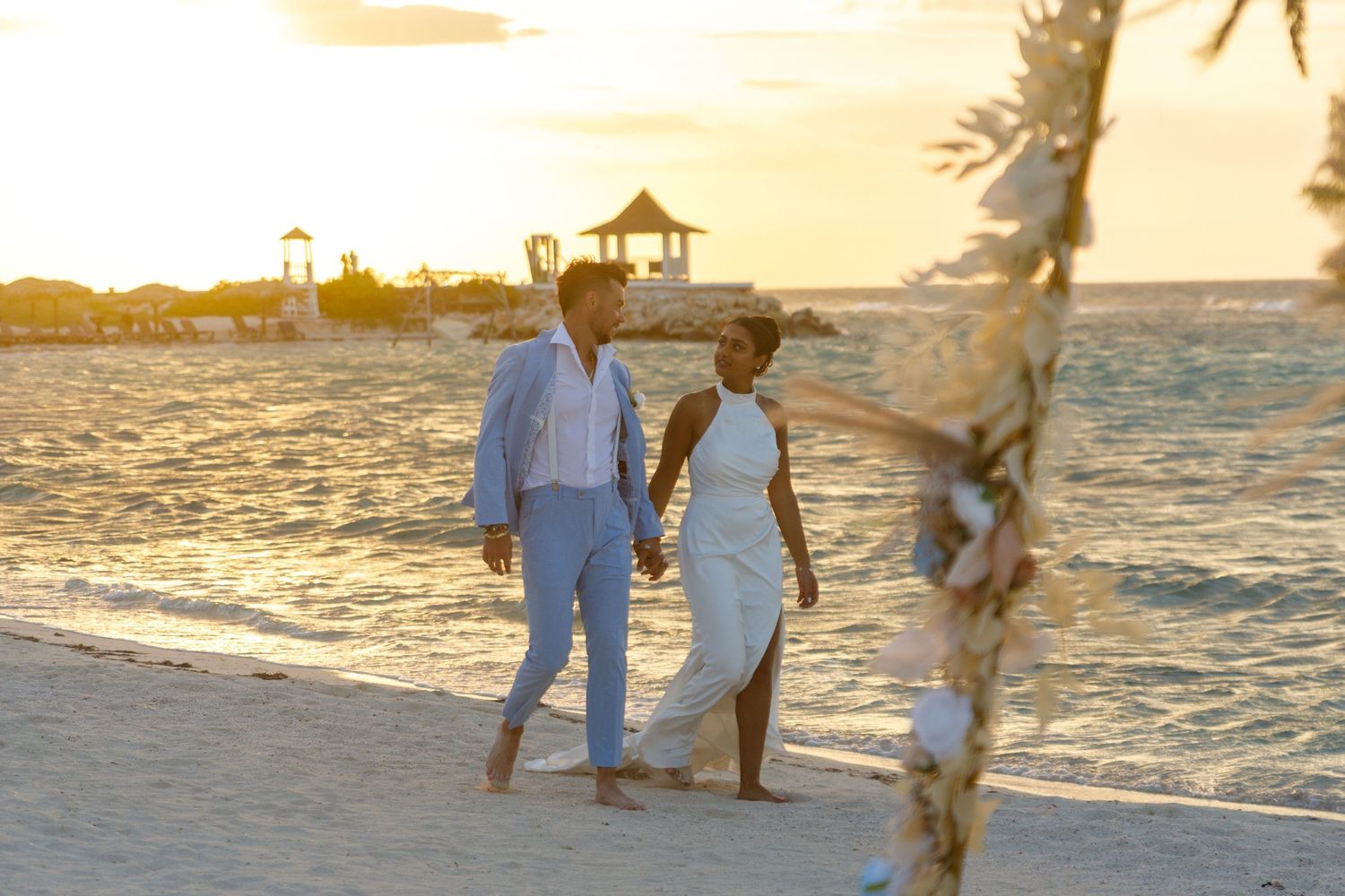 Couple holding hands walking on a beach at sunset during a romantic destination wedding ceremony.