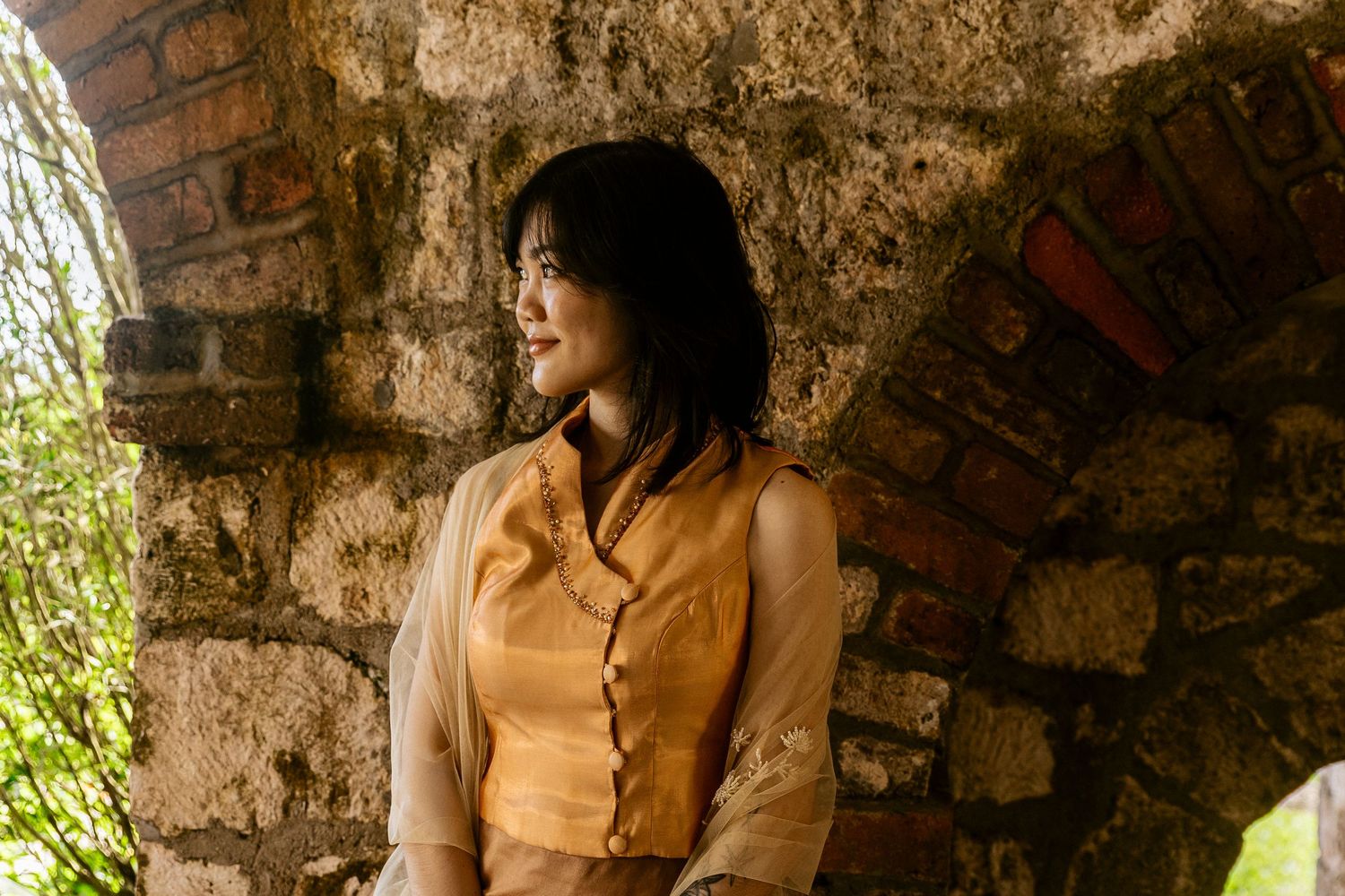 Young Asian woman in elegant orange dress leaning against a rustic stone arch wall outdoors.