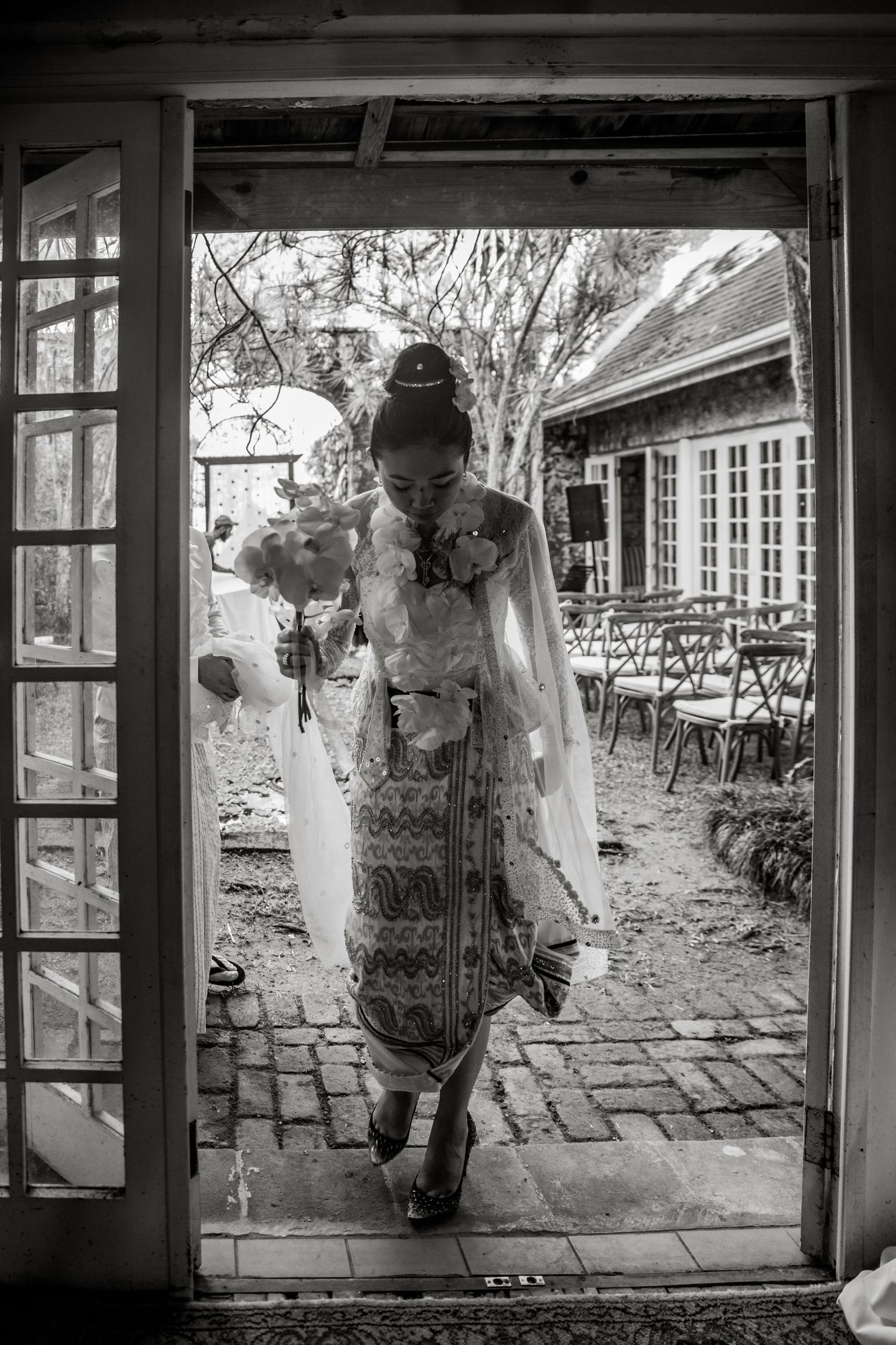 Black and white photo of bride in traditional Myanmar dress walking through French doors.