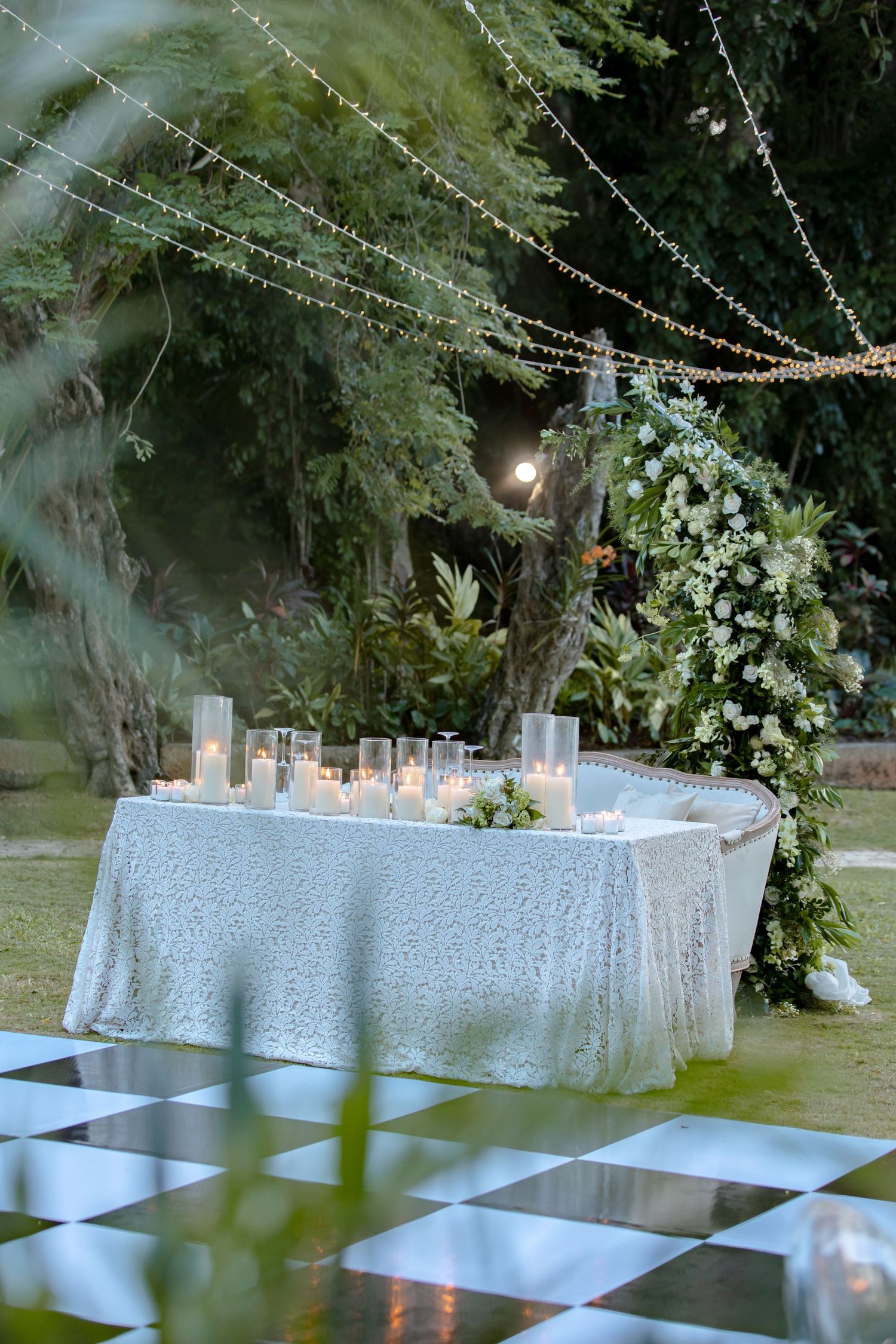 Elegant outdoor wedding reception table with white sequin linen, candles, floral arch, fairy lights, and checkered dance floor.