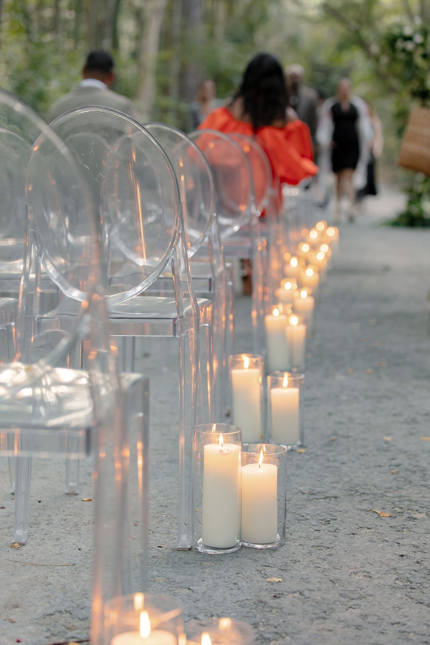 Lit white pillar candles lining wedding ceremony aisle beside transparent ghost chairs in forest setting.